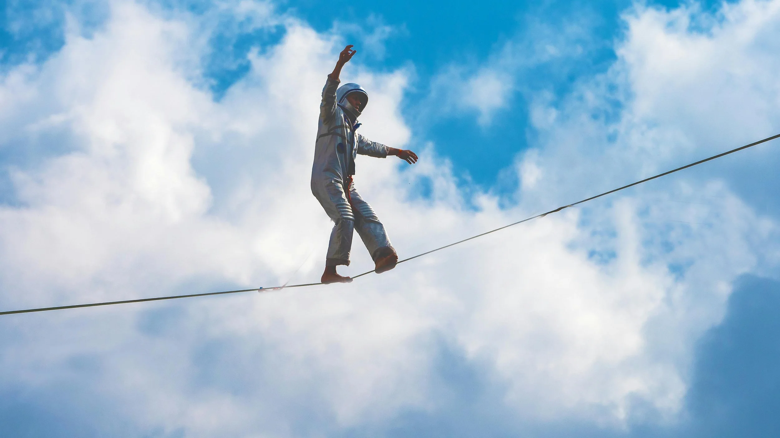 A person walking a tightrope against a bright blue cloudy sky, arms outstretched for balance, conveying the careful judgment required between speed and integrity.