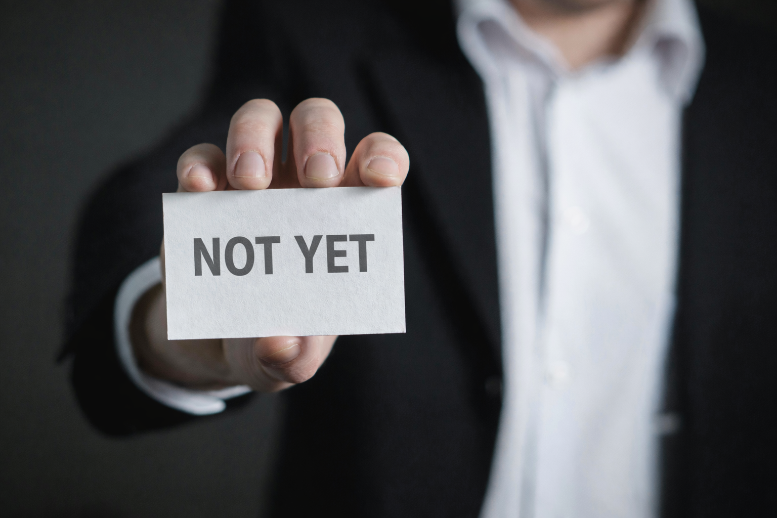 A person in a dark suit holding a white card that reads “NOT YET,” symbolizing deliberate timing and strategic restraint in decision-making.