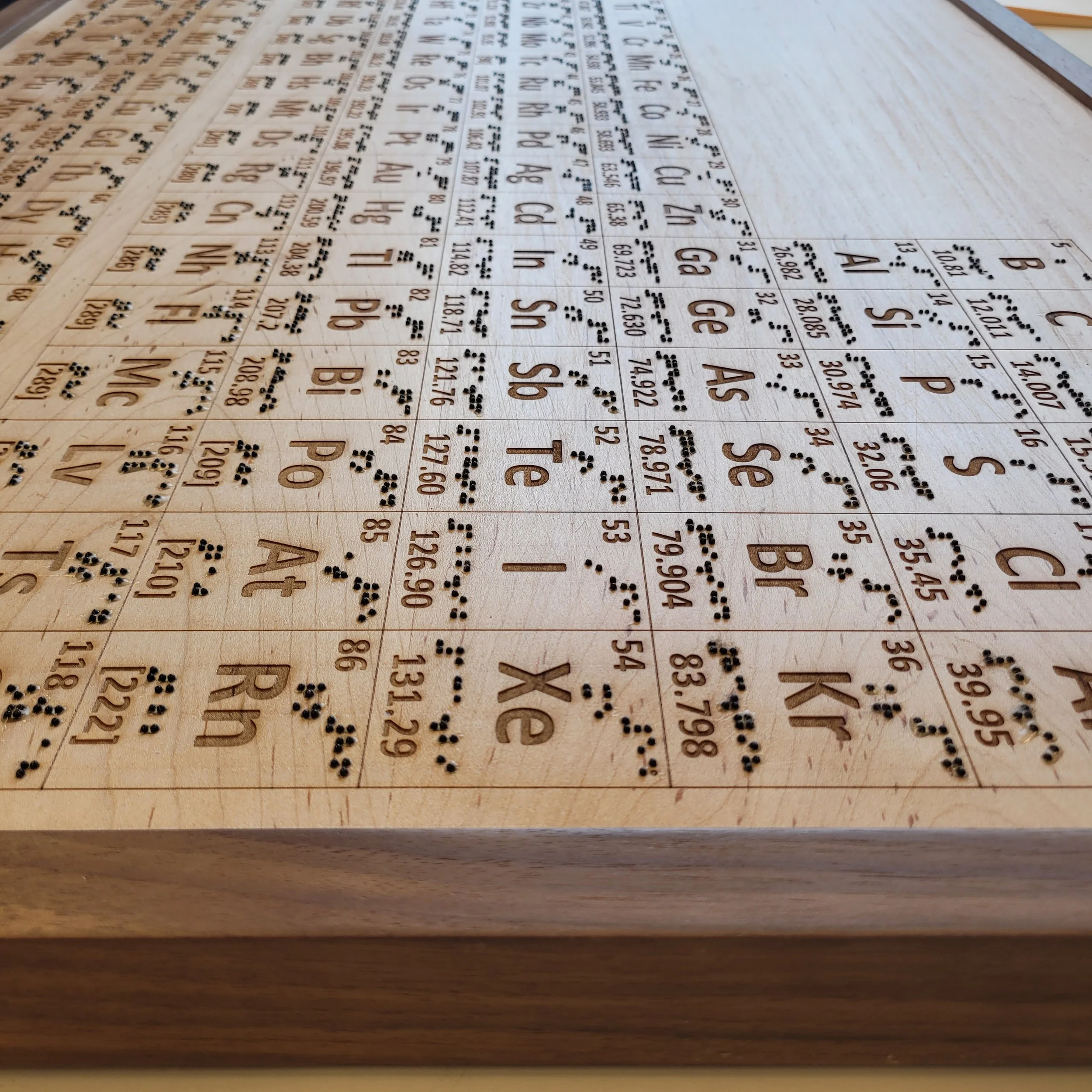 An angled overhead view of the complete wooden tactile periodic table set in a dark walnut frame, showing the full grid of laser-engraved element cells with Braille dots across the entire surface.