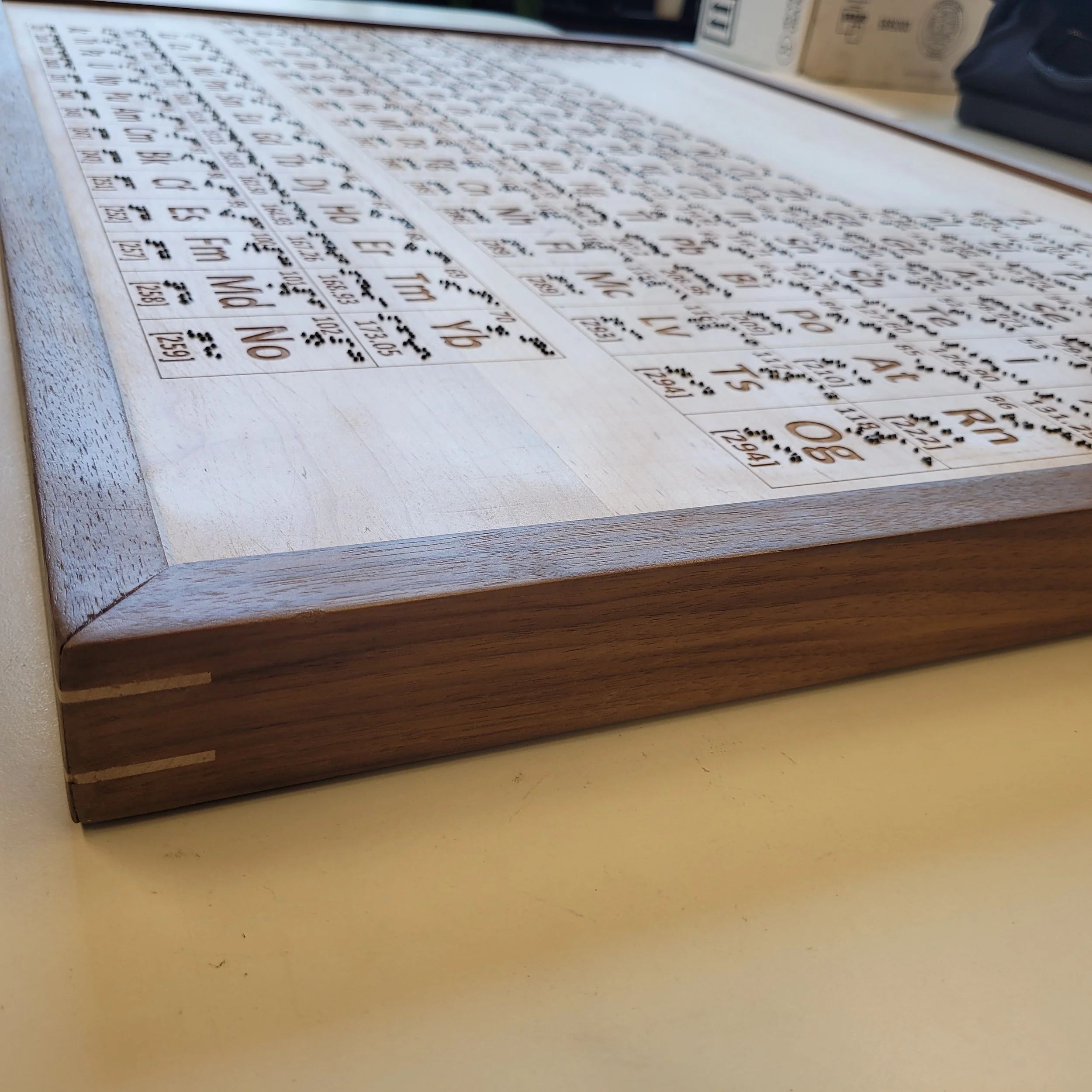 A low-angle side profile shot of the wooden tactile periodic table, highlighting the thickness of the maple board and the dark walnut wooden frame construction underneath.