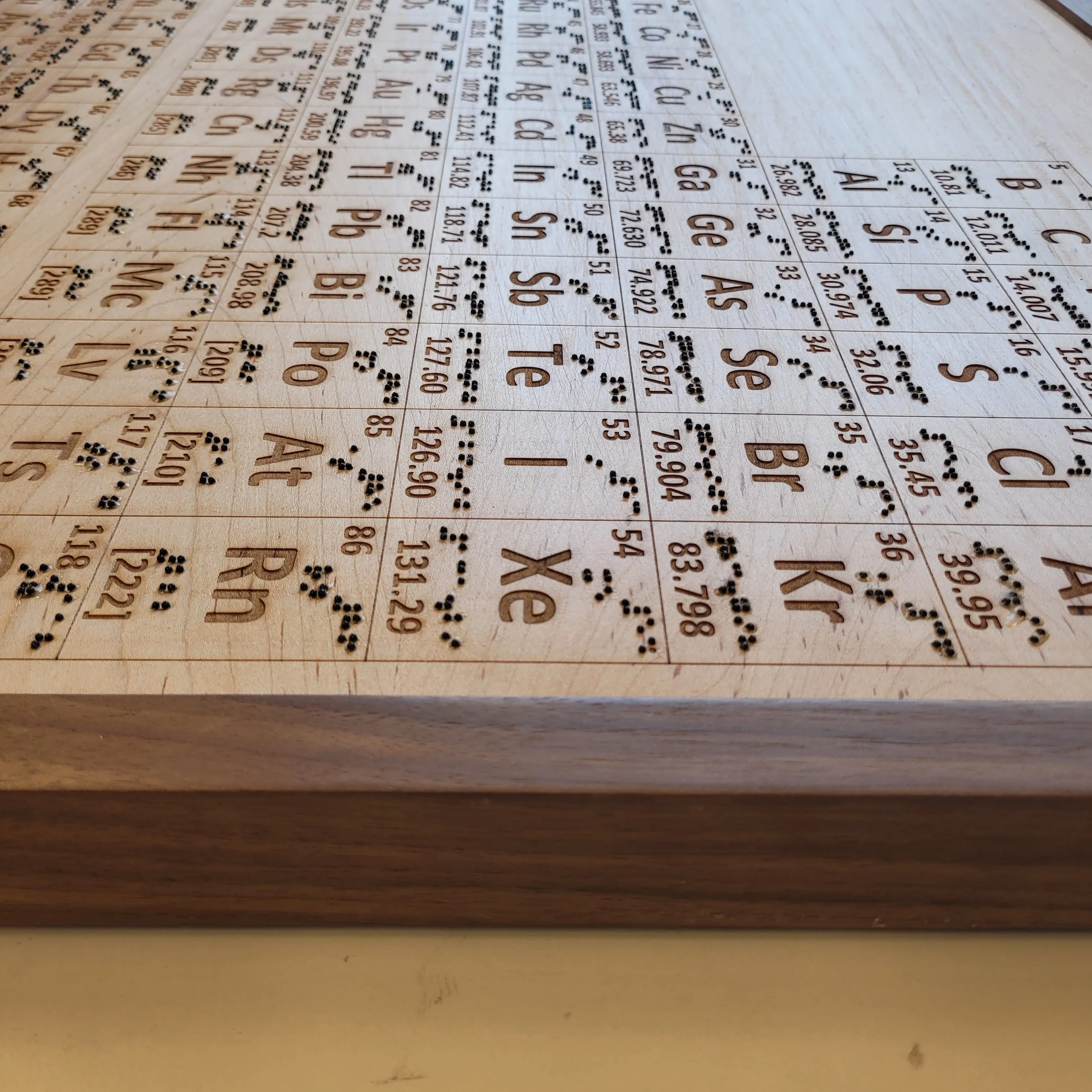 A close-up angled view of the top edge and corner of the wooden tactile periodic table, showing the laser-engraved element cells and the dark wooden border frame from the side.