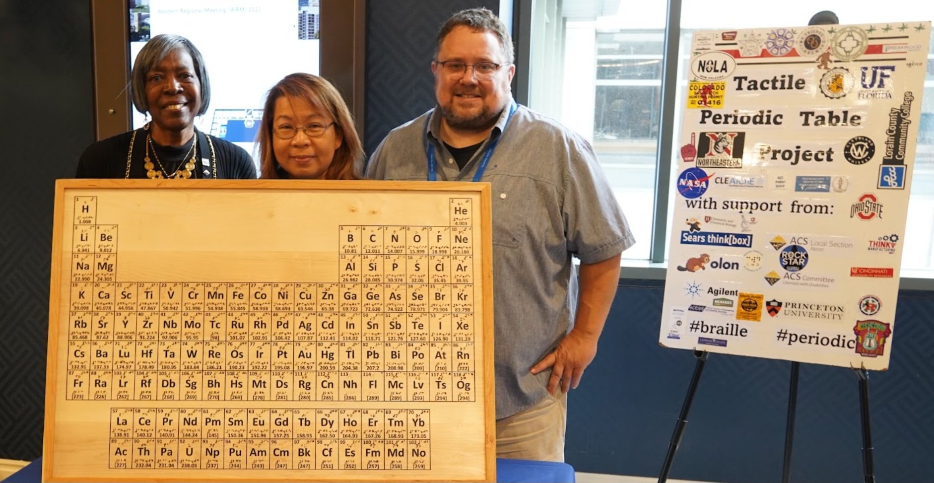 Three people smiling behind a large wooden tactile periodic table with Braille markings, next to a poster reading "Tactile Periodic Table Project" with sponsor logos, at a conference event.