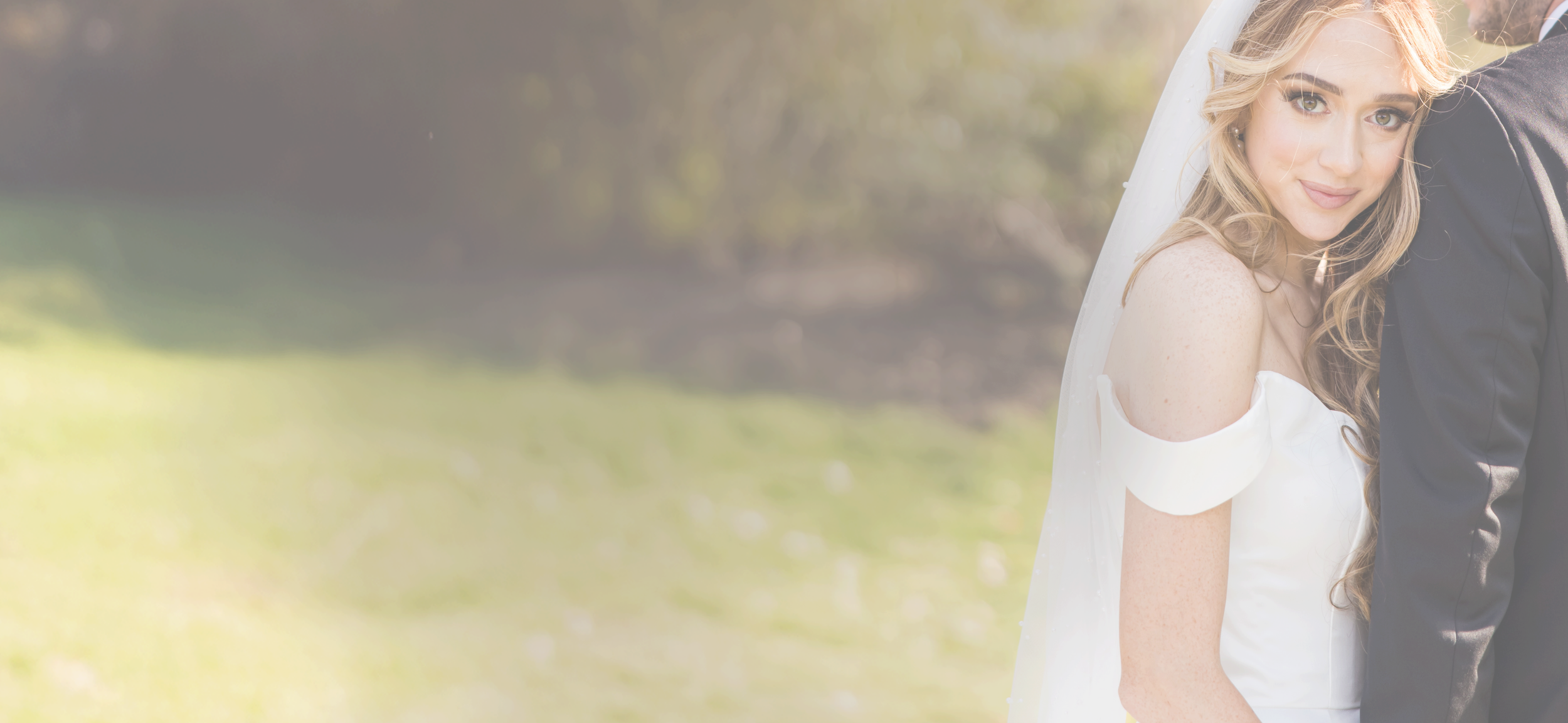 A bride with wavy blonde hair in a white wedding dress is smiling at the camera, standing outdoors with a groom in a dark suit beside her. The bride has a veil and is in a natural setting with sunlight and blurred greenery in the background.