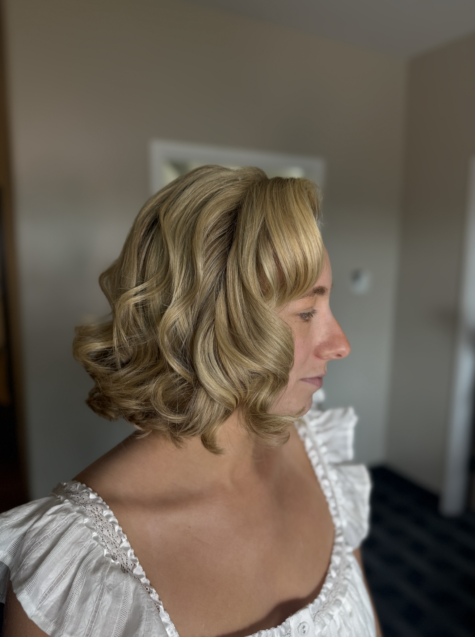 A woman with styled blonde hair in loose curls, wearing a white top with ruffled sleeves.
