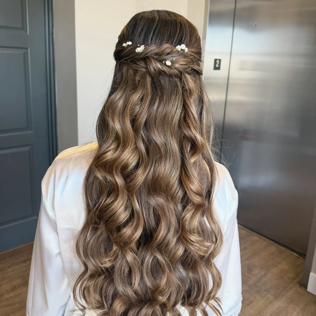 Back view of a woman with long, wavy, light brown hair styled in loose curls, adorned with small white flower hair clips.
