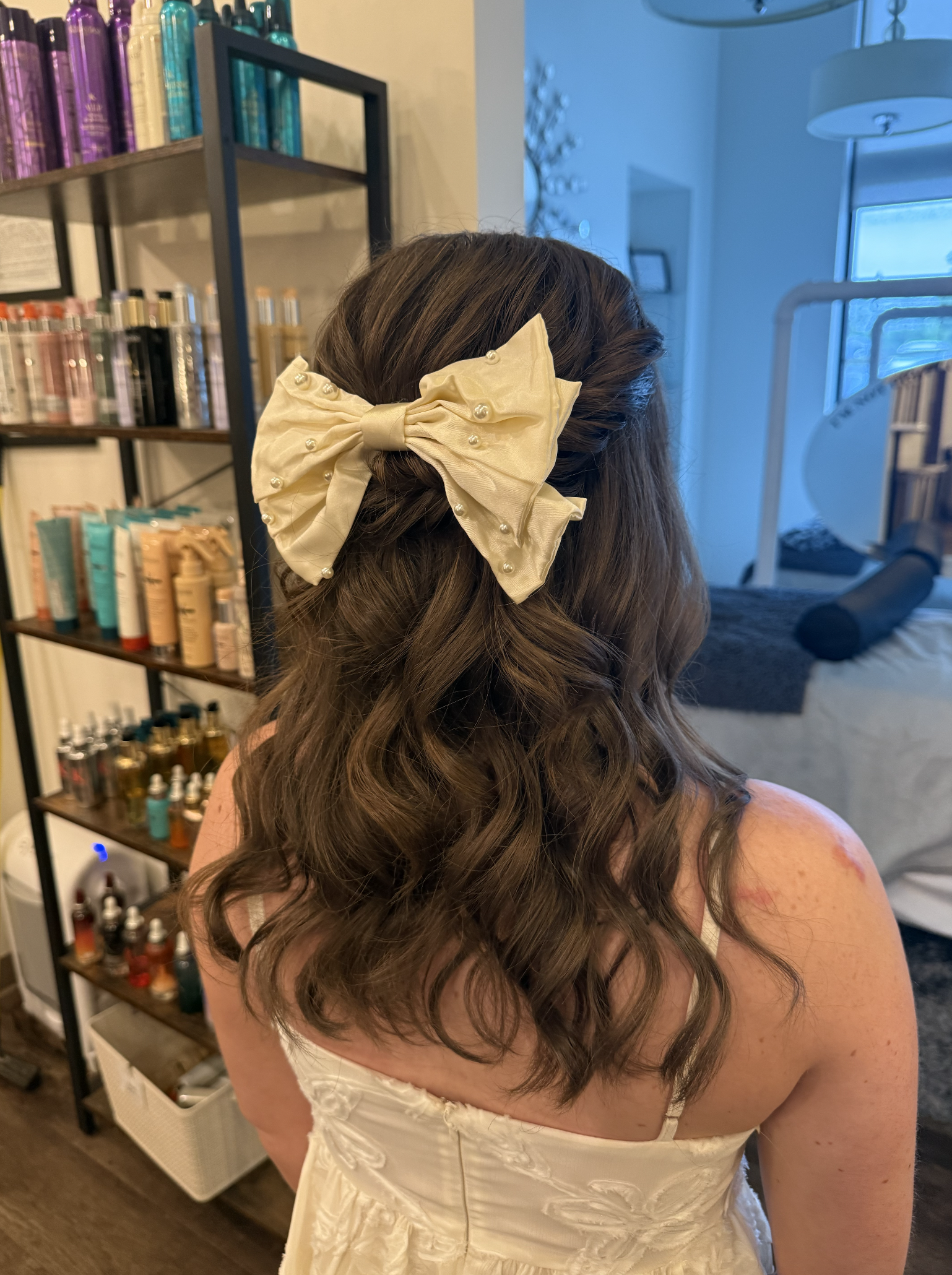 Back view of a woman with long, wavy brown hair wearing a white dress and a large, cream-colored bow hair accessory decorated with small pearls.