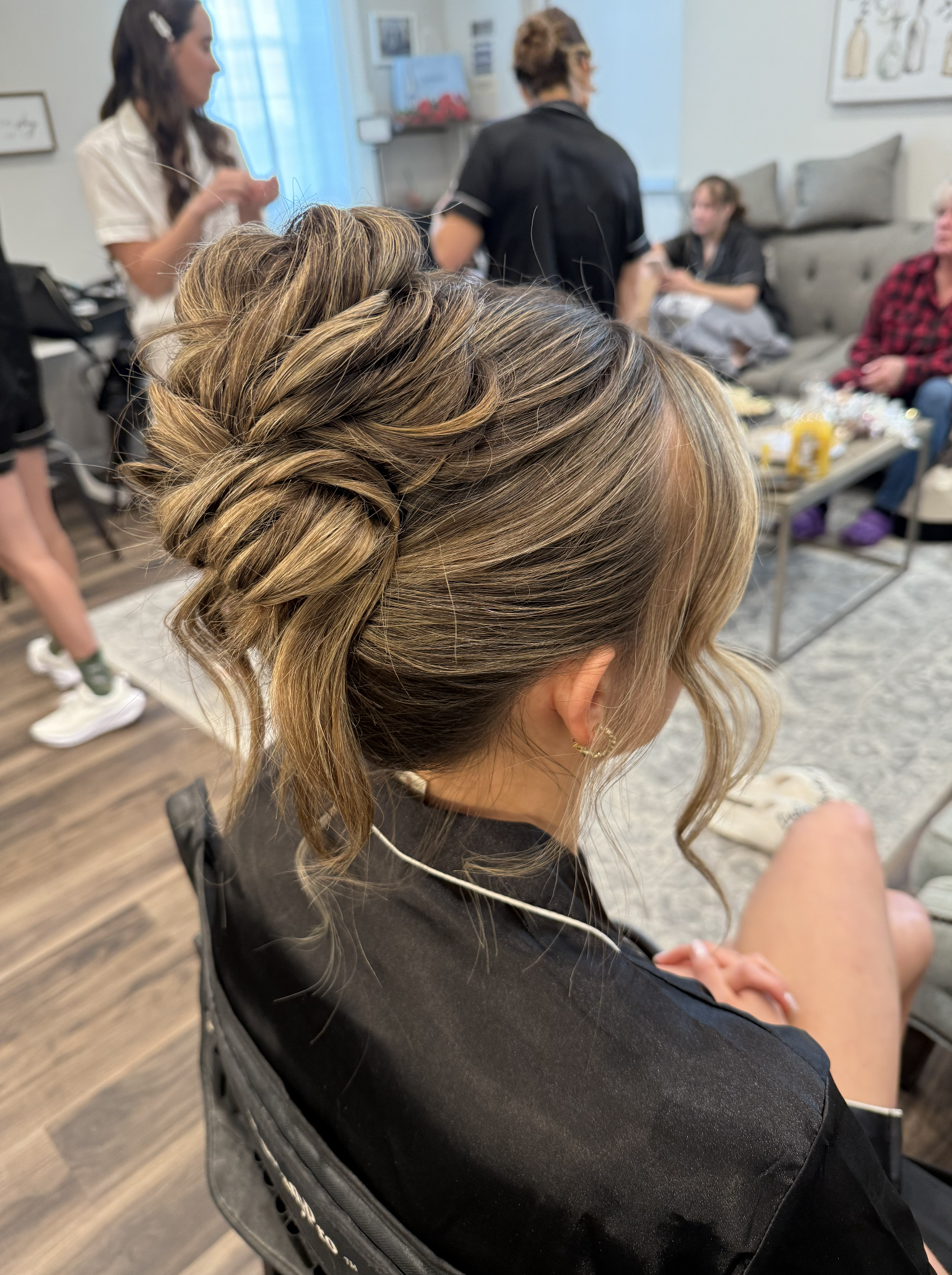 A woman with styled, braided hair sitting in a hair salon, surrounded by people, including a woman in a white shirt, a man in black, and others sitting on a couch.