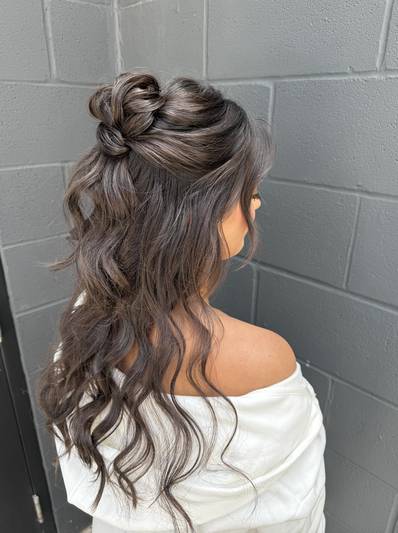 Side view of a woman with long, wavy brown hair styled with a twisted half-updo, standing against a gray brick wall.