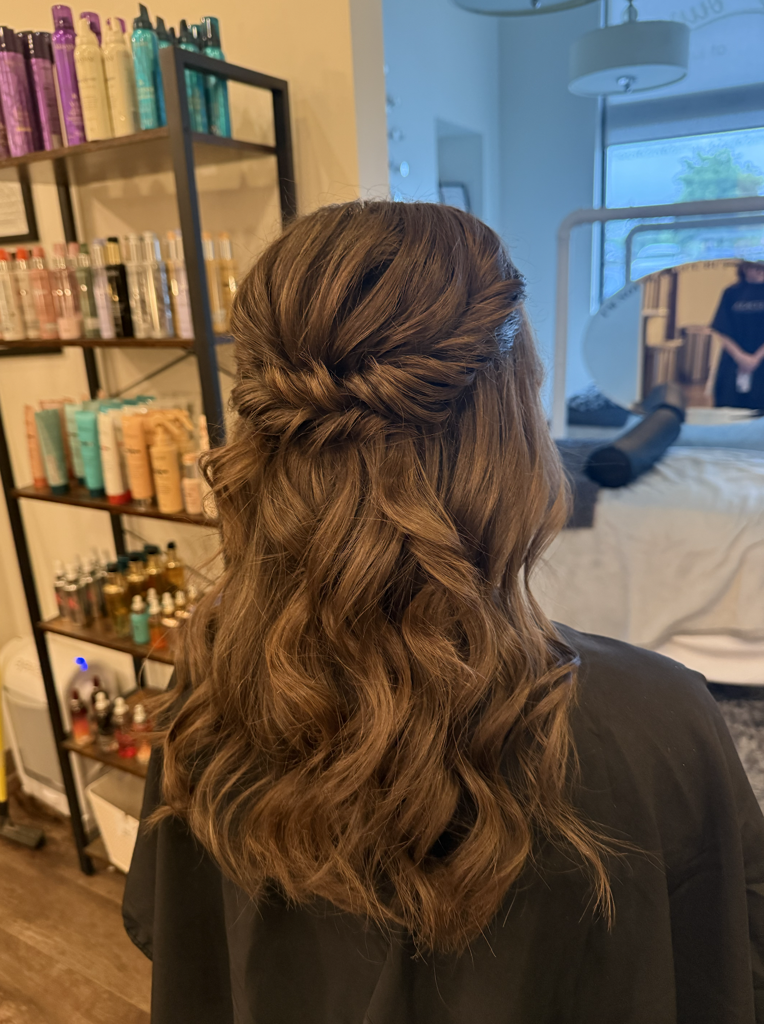 Back of a woman's head showing styled, wavy brown hair with a twist at the top, in a salon. Shelves with hair products are in the background, along with a salon mirror reflecting a person.