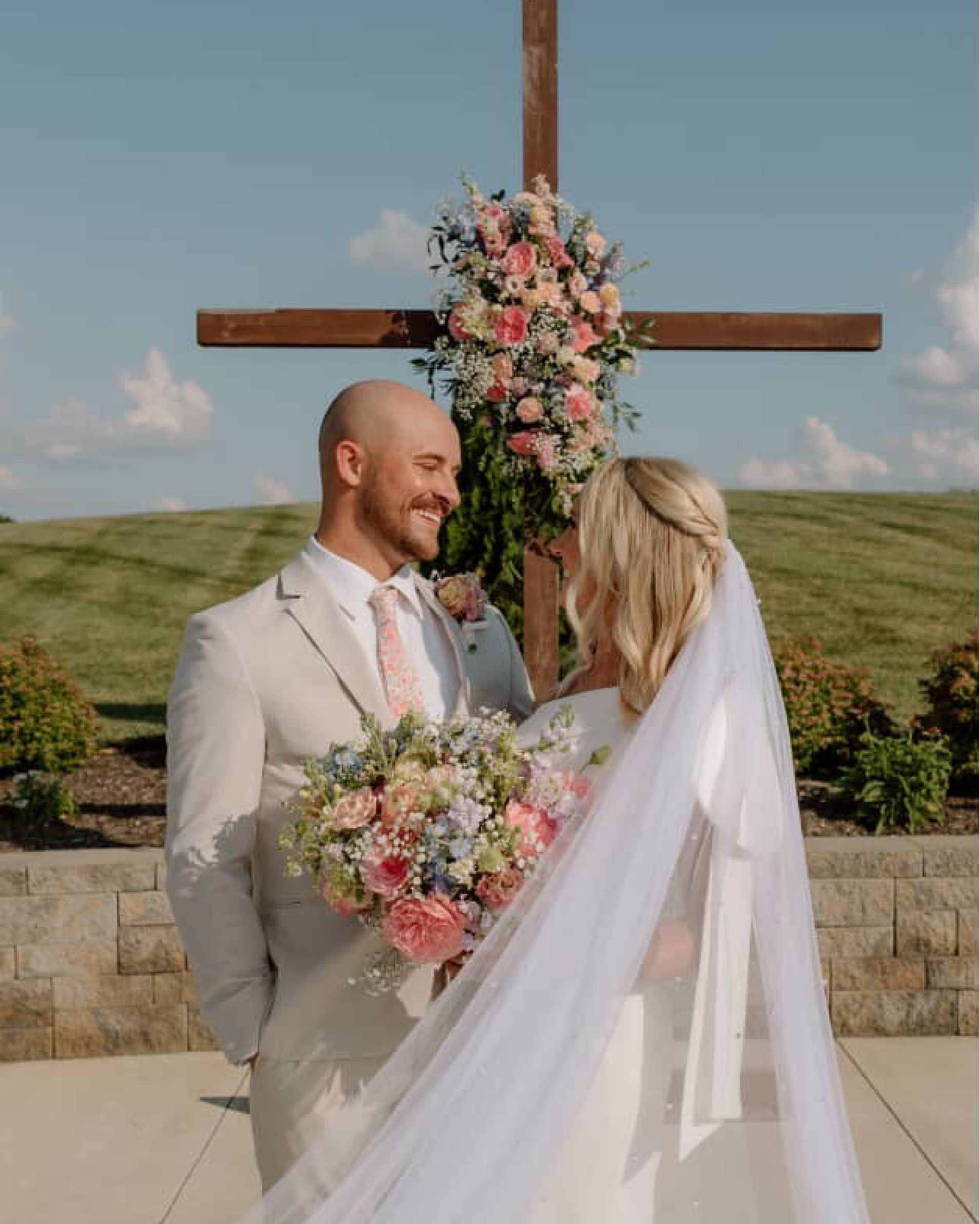 A bride and groom standing outdoors, smiling at each other, during their wedding ceremony with a wooden cross decorated with pink, white, and purple flowers in the background.