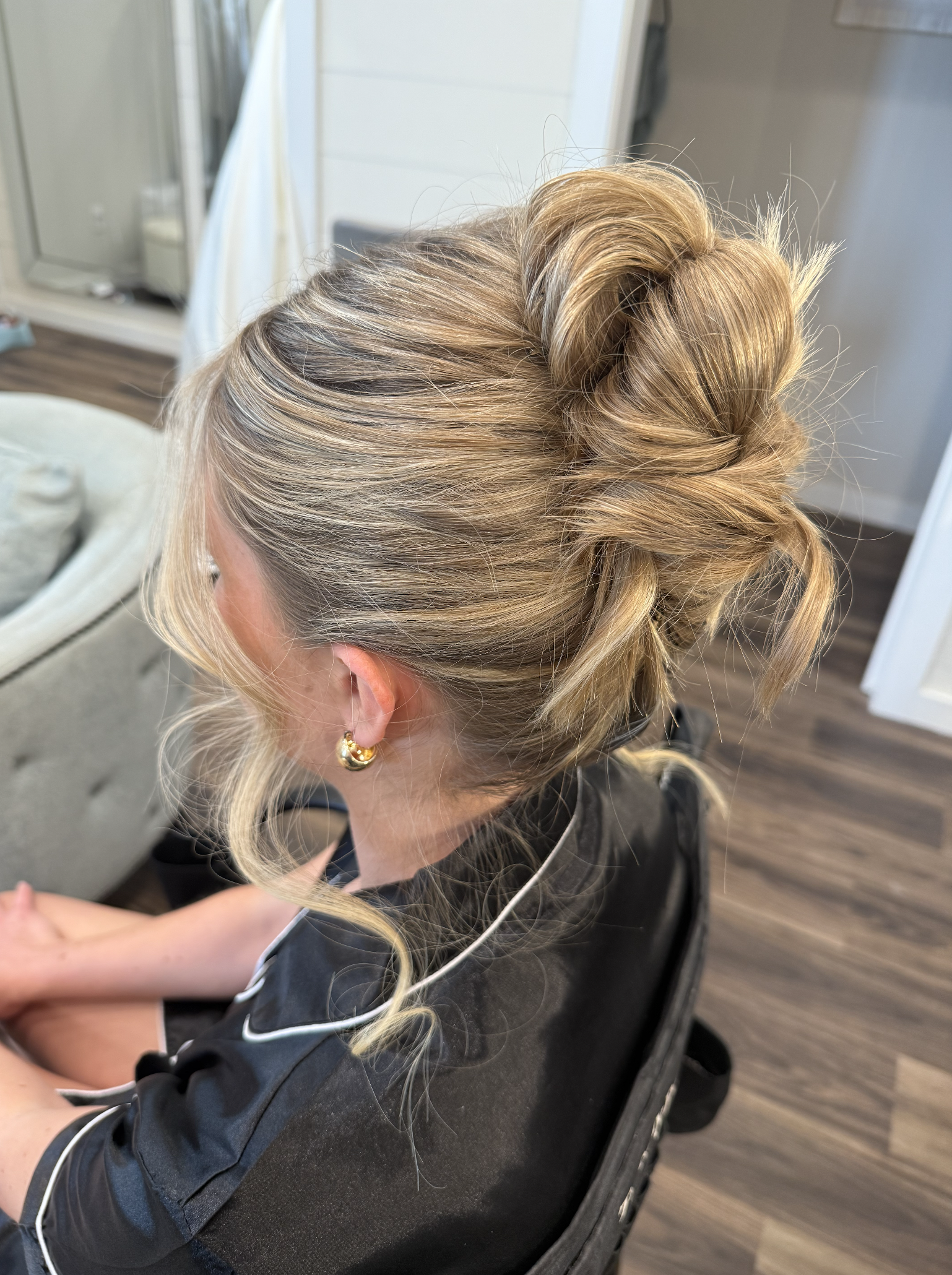 A woman with blonde hair styled in an elegant updo, sitting in a salon chair, wearing earrings and a black top with white piping.