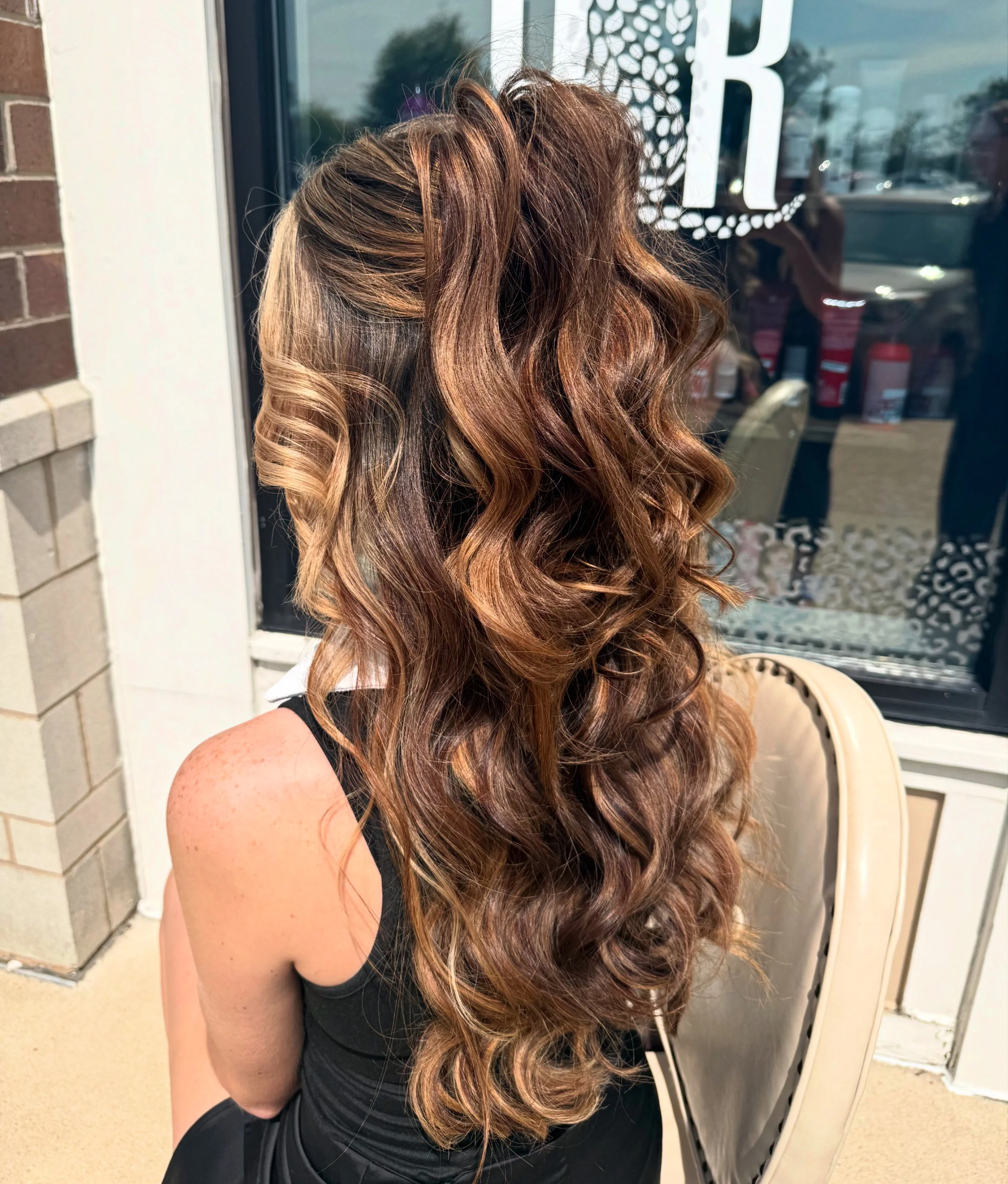 A woman with long, wavy brown hair sitting outdoors near a glass door.