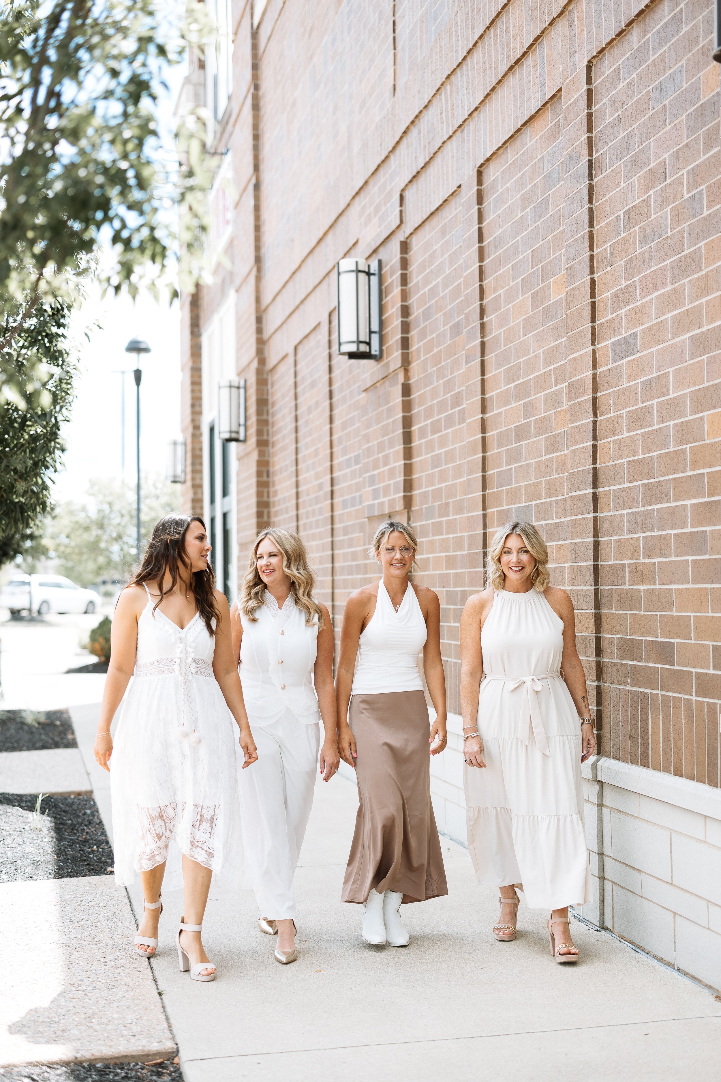 Four women dressed in white and beige walking along a sidewalk past a brick building, smiling and chatting