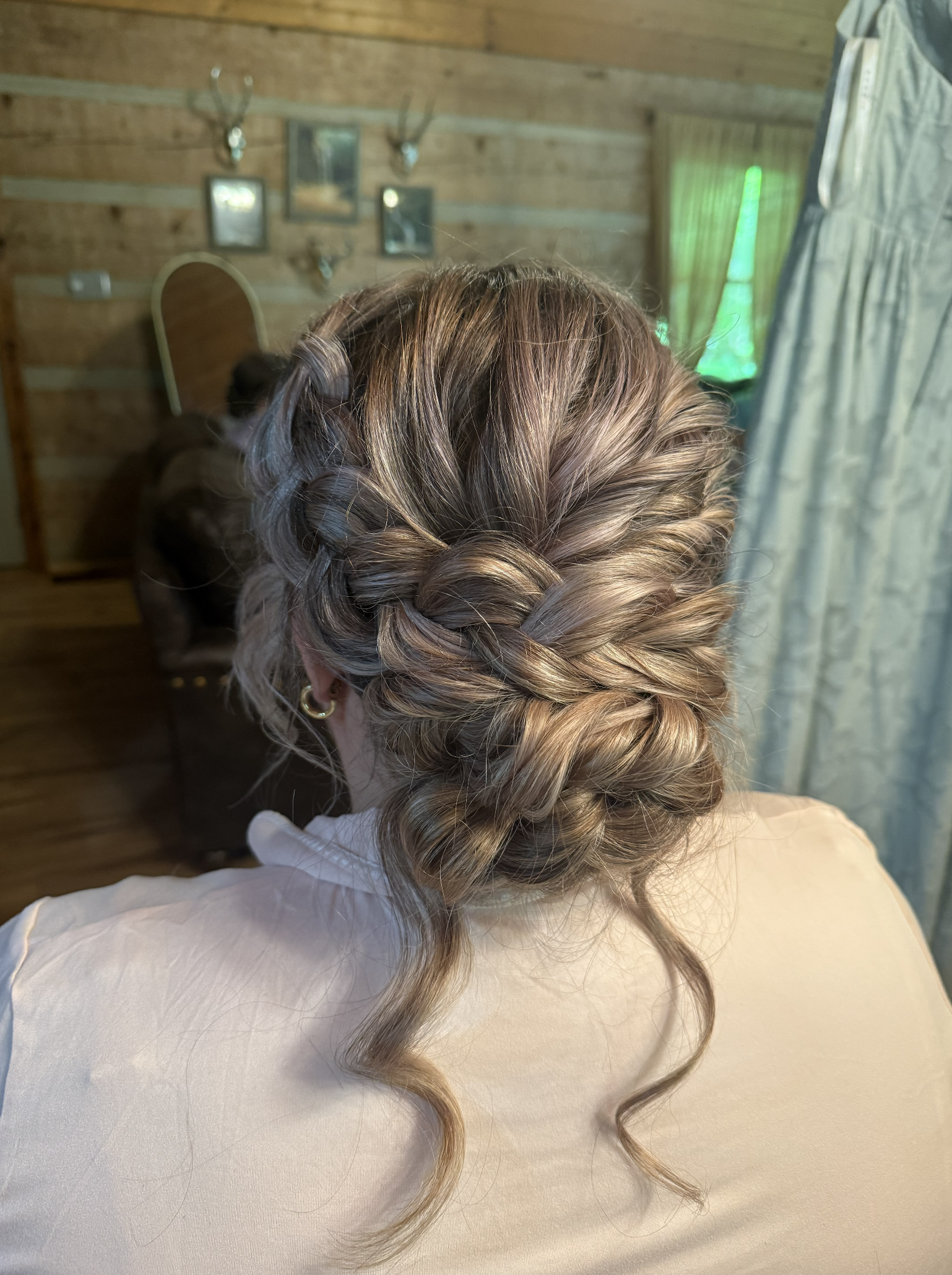 Back of a woman with styled blonde hair in an intricate braided updo with loose curls, wearing a beige outfit in a rustic wooden room.