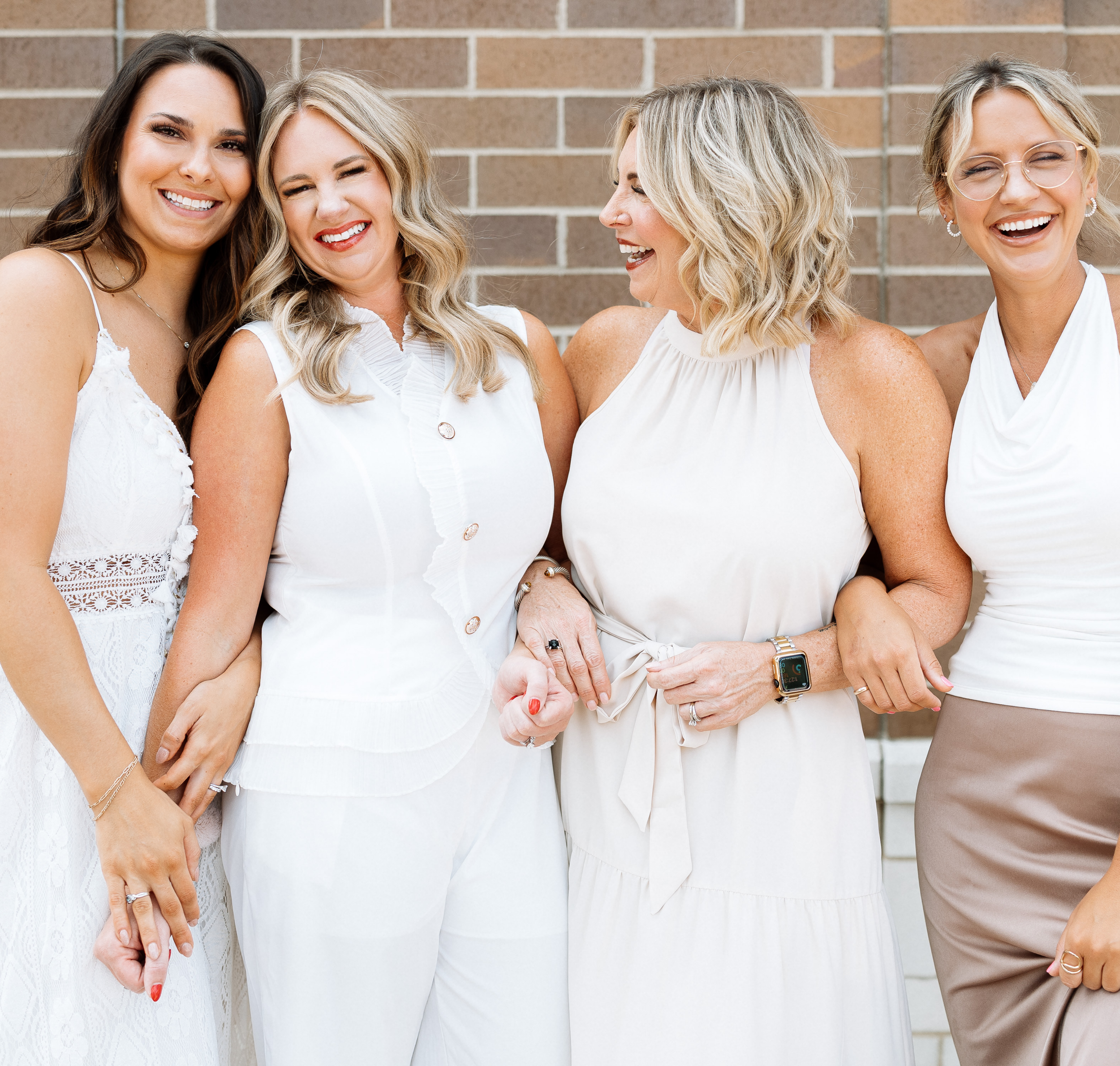 Four women in white attire, smiling and laughing together against a brick wall.