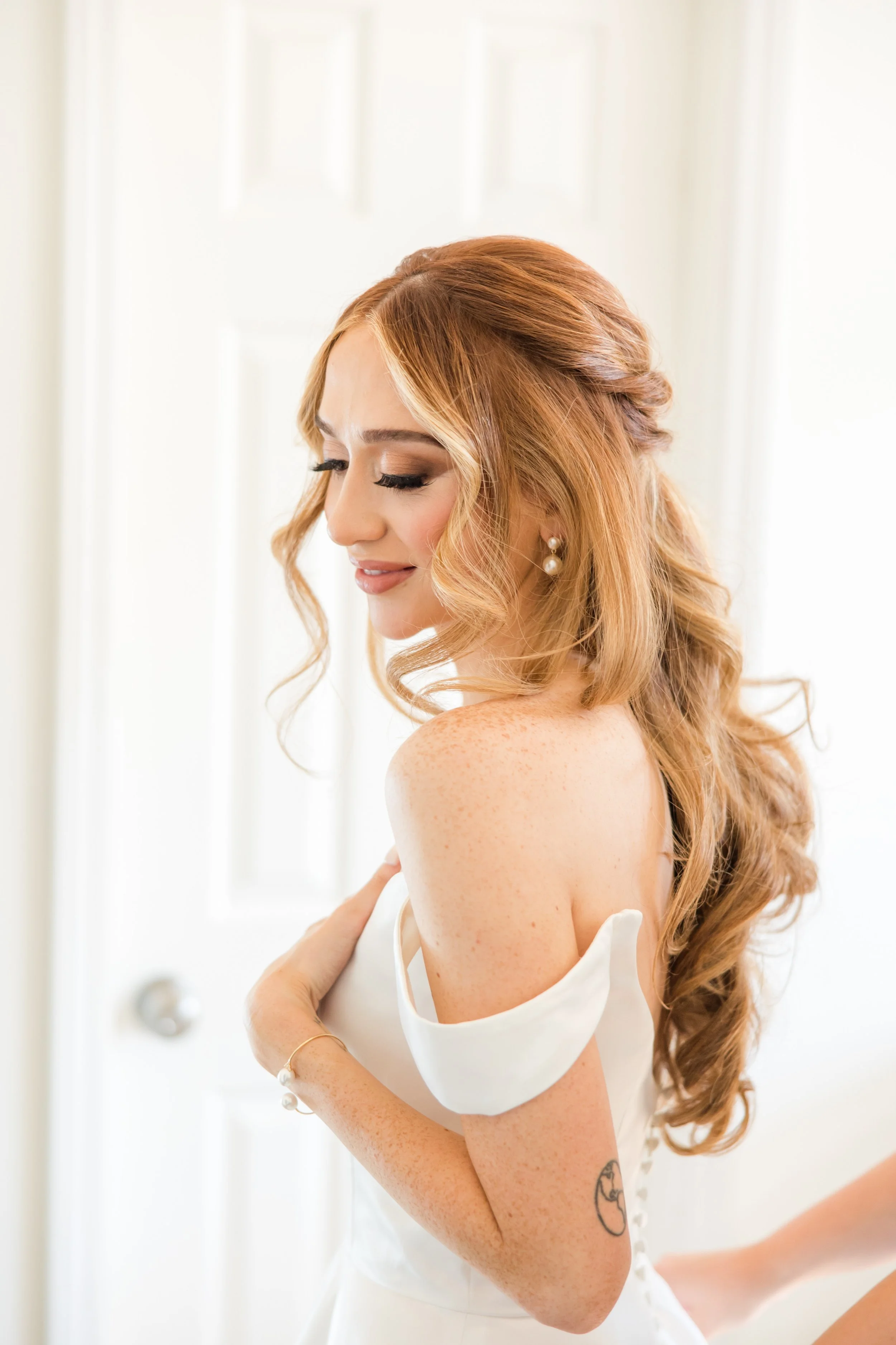 A woman with long wavy red hair, tan freckles, and pearl earrings is wearing a white off-shoulder dress, smiling softly with her eyes closed.