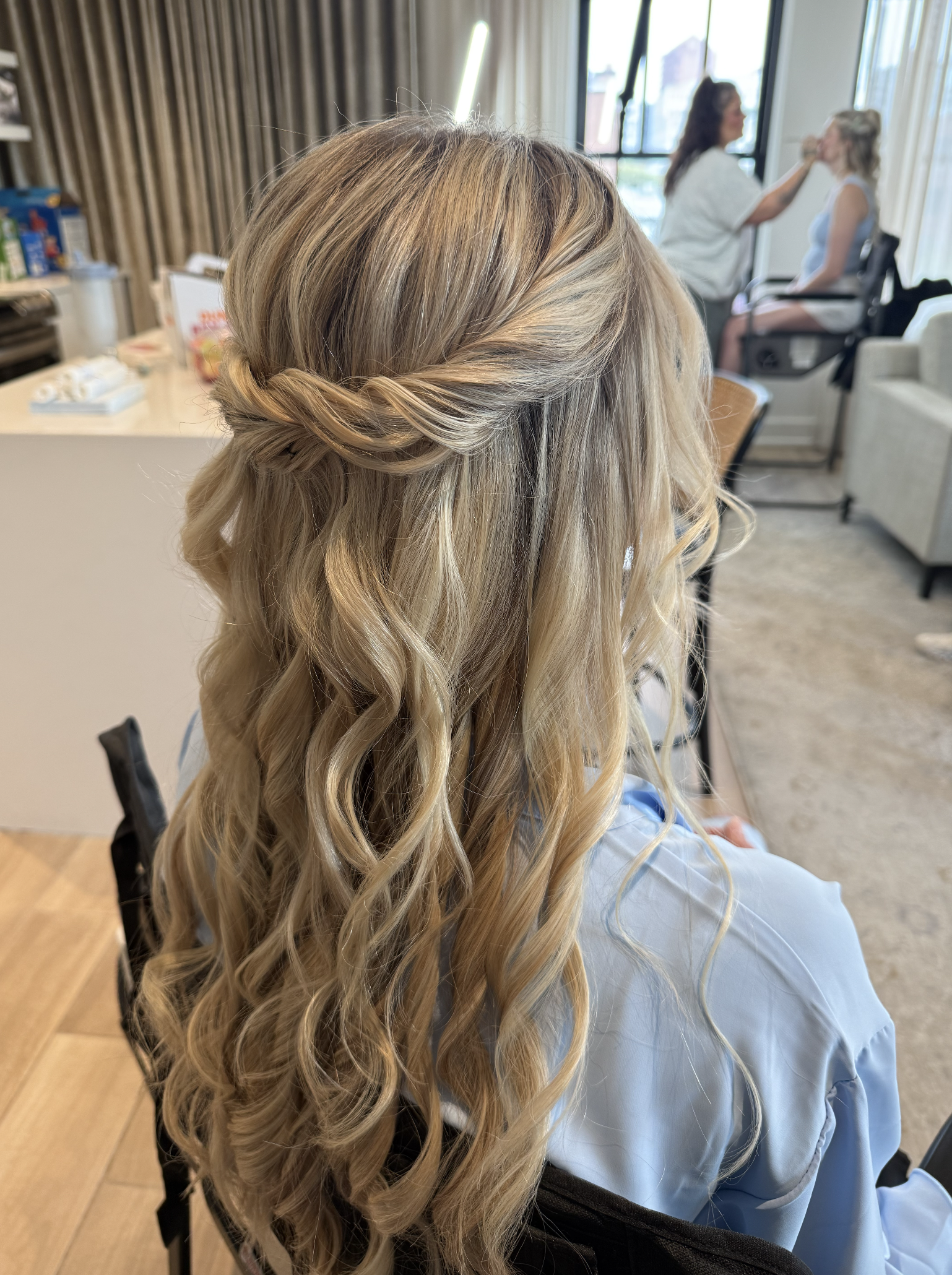 Woman with long blonde, curly hair styled with a twisted braid on the side, sitting in a salon chair.