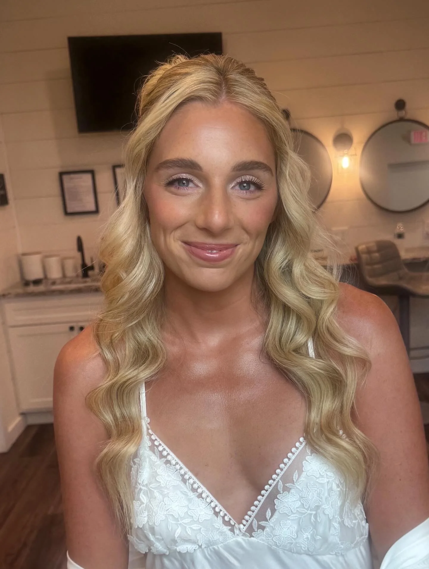 A woman with long blonde wavy hair, wearing a white lace dress, smiling in a room with wood-paneled walls, round mirrors, and a television.