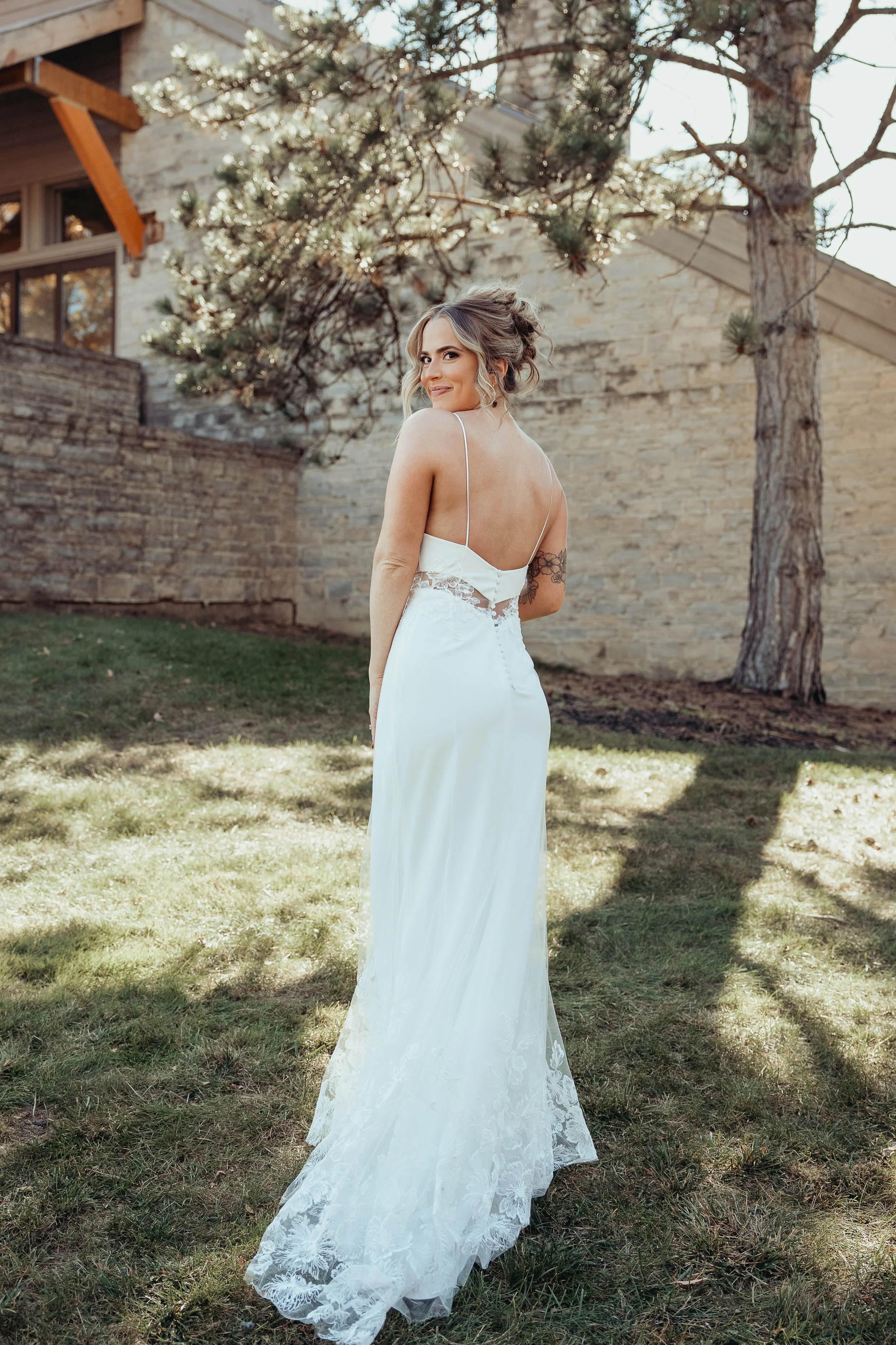 A woman in a white wedding dress standing on grass outside near trees and a stone building, looking over her shoulder.