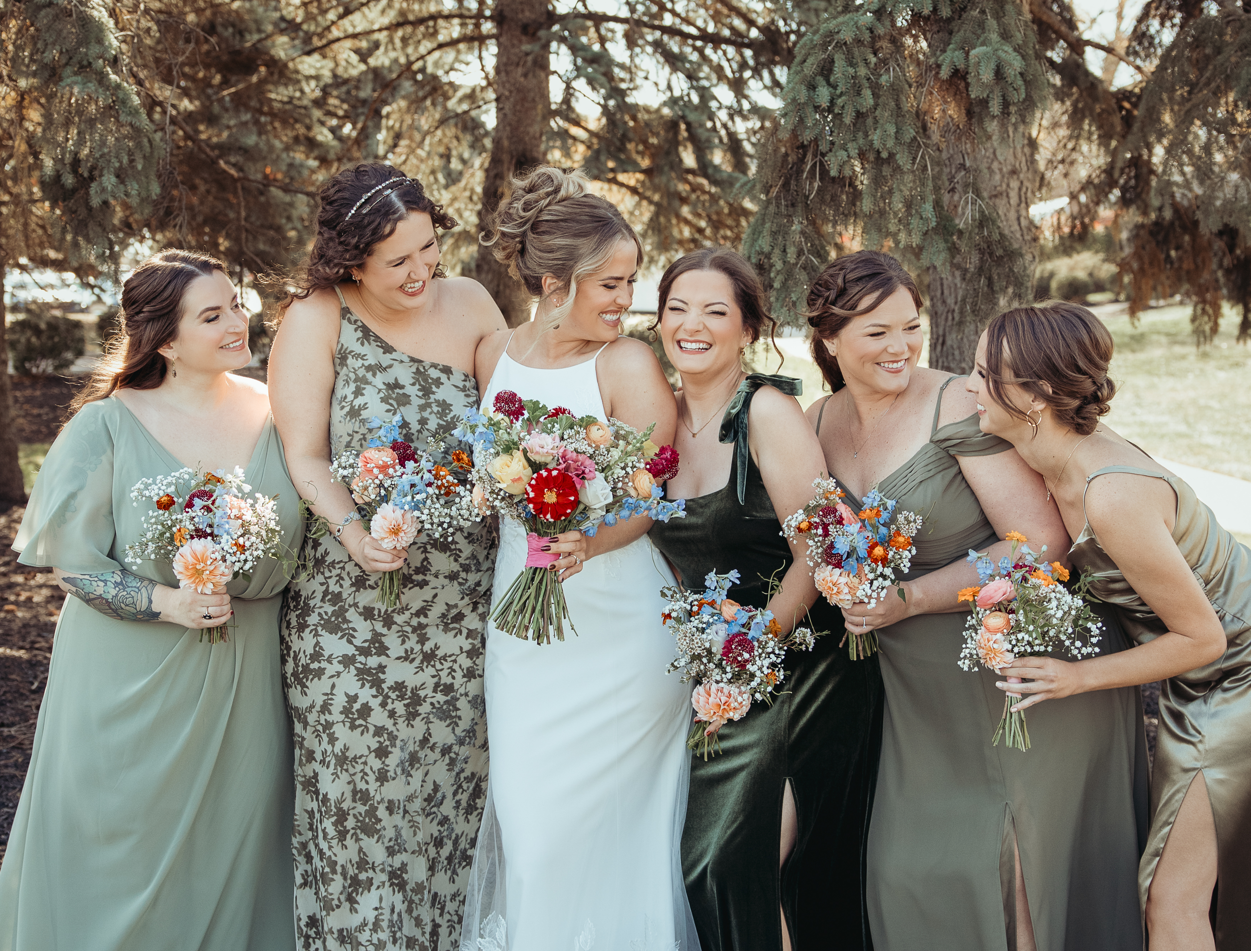 A group of seven women dressed in formal attire, smiling and laughing together outdoors near a large tree, with some holding bouquets of flowers. The woman in the center is wearing a white dress and holding a bouquet, suggesting she might be the bride.
