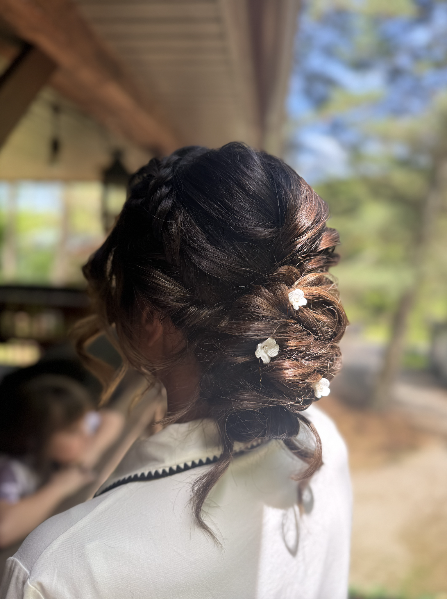 A woman with dark brown hair styled in an elegant updo decorated with small white flower hairpins, standing outdoors near a wooden structure and trees, with another person blurred in the background.