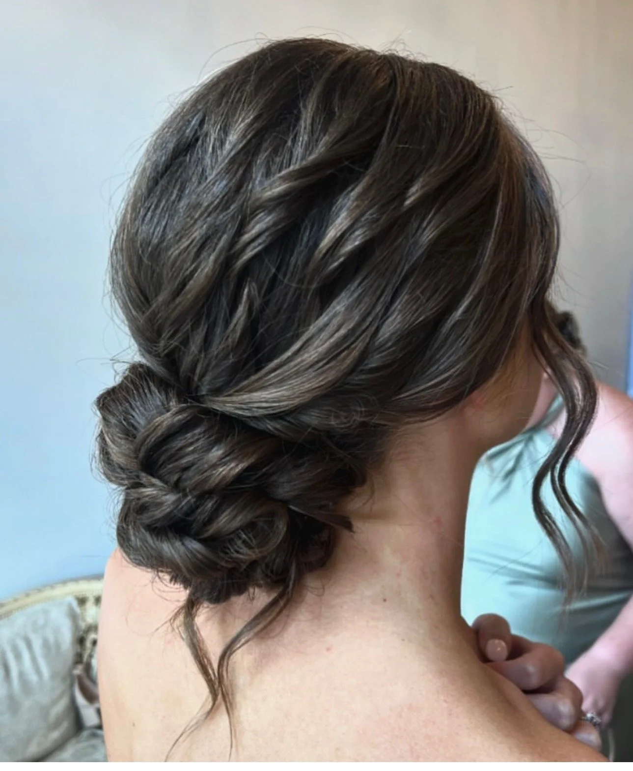 Side view of a woman with a wavy updo hairstyle with a braid, her skin visible, and a curled strand hanging loosely, sitting indoors near a light-colored wall.