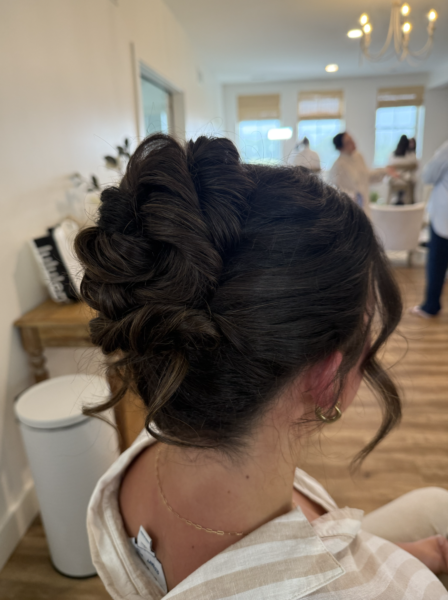A woman with dark hair styled in an elegant updo, with curly loose strands, sitting indoors in a light-colored room with wood floors, and friends or family in the background.