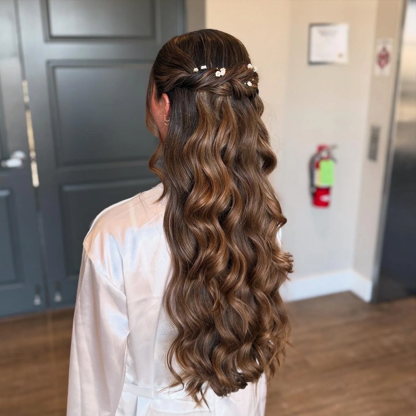 Back view of a woman with long, wavy brown hair decorated with small white flowers, standing inside a room with gray door and fire extinguisher in the background.