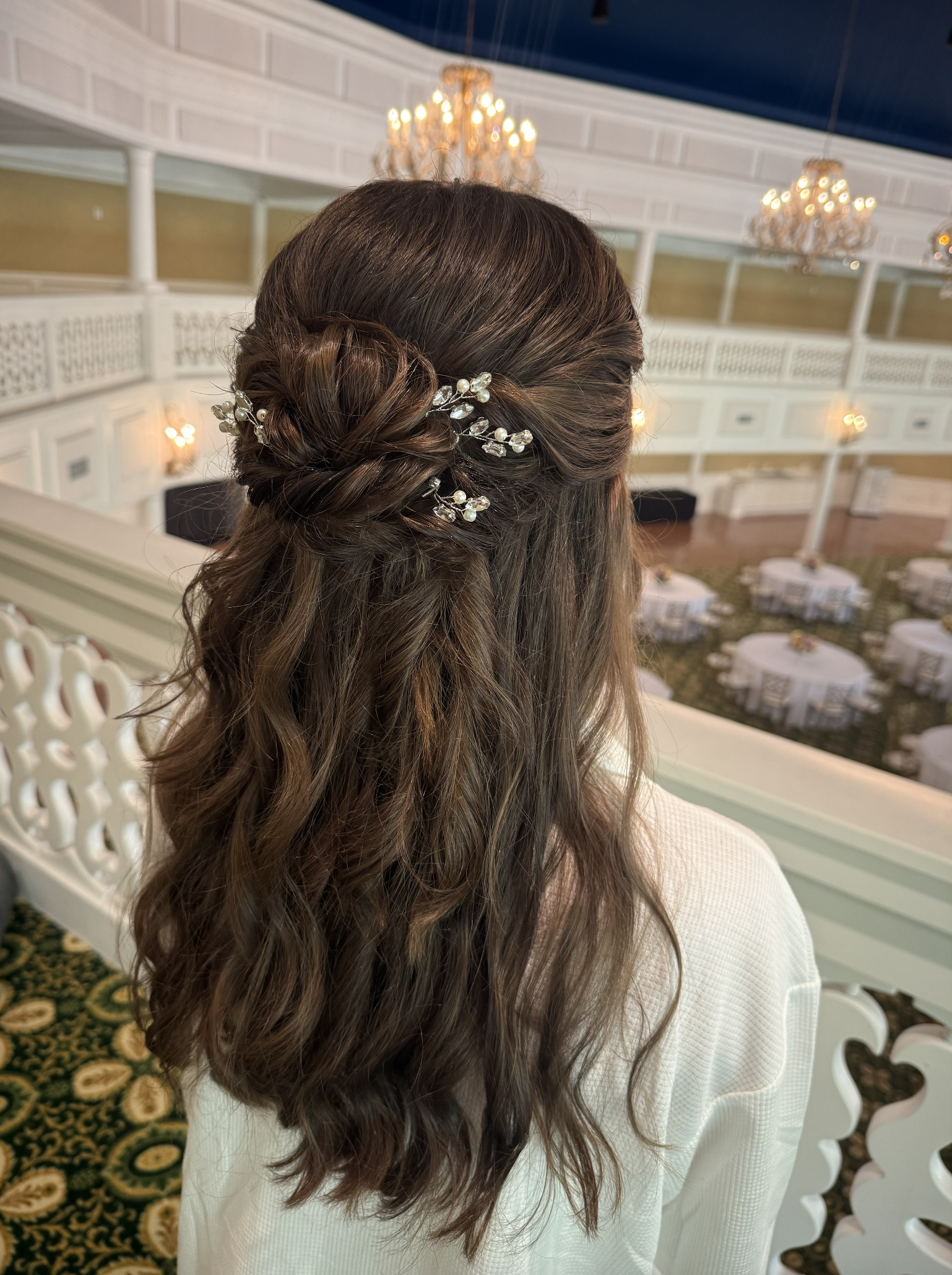 Back of a woman's head with long wavy brown hair styled with a braid and decorative pearl and pearl-studded hairpiece, at an indoor event venue with chandeliers and round tables.