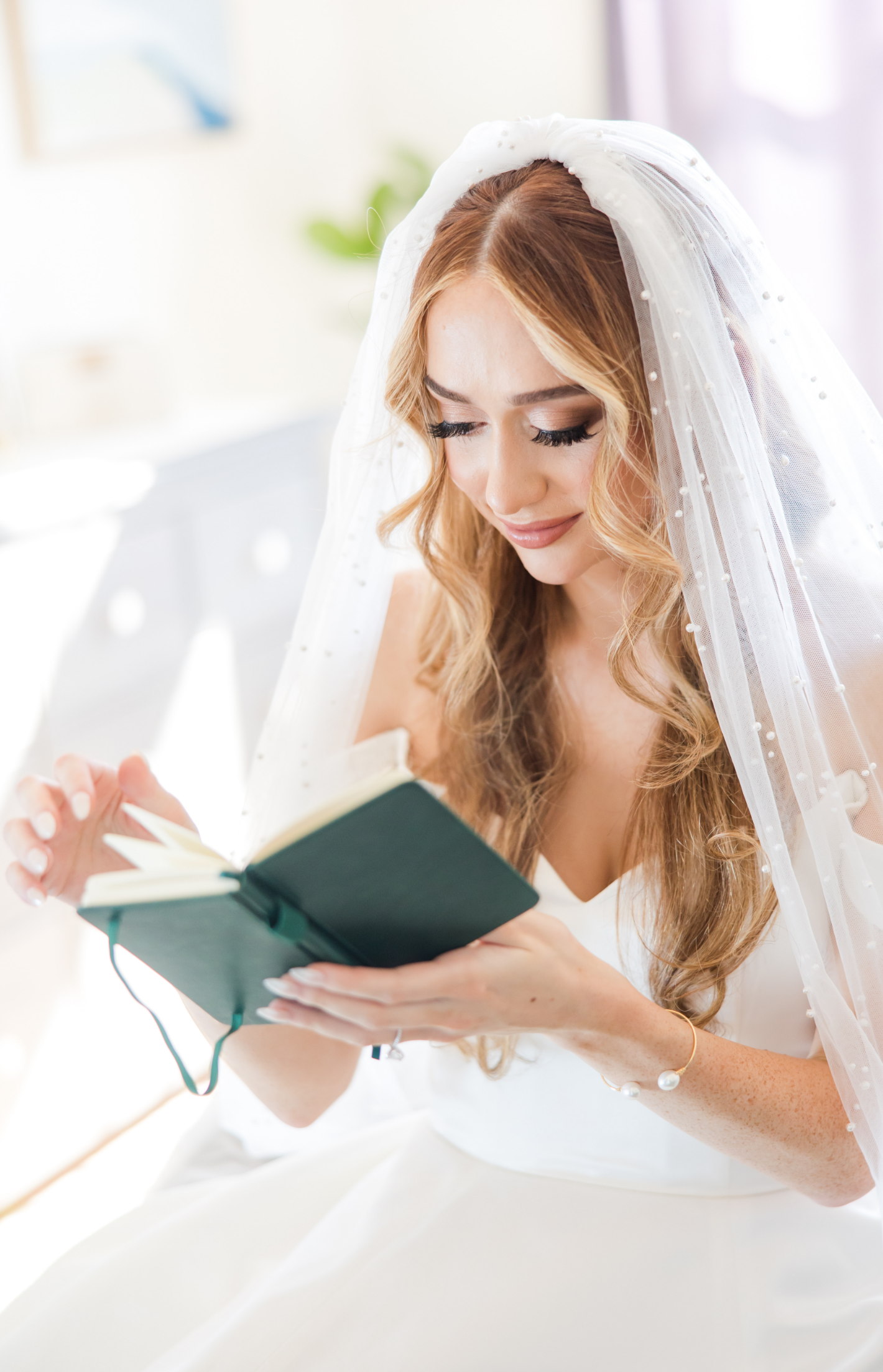 A bride with long curly hair wearing a veil and white dress, reading a small book or journal.