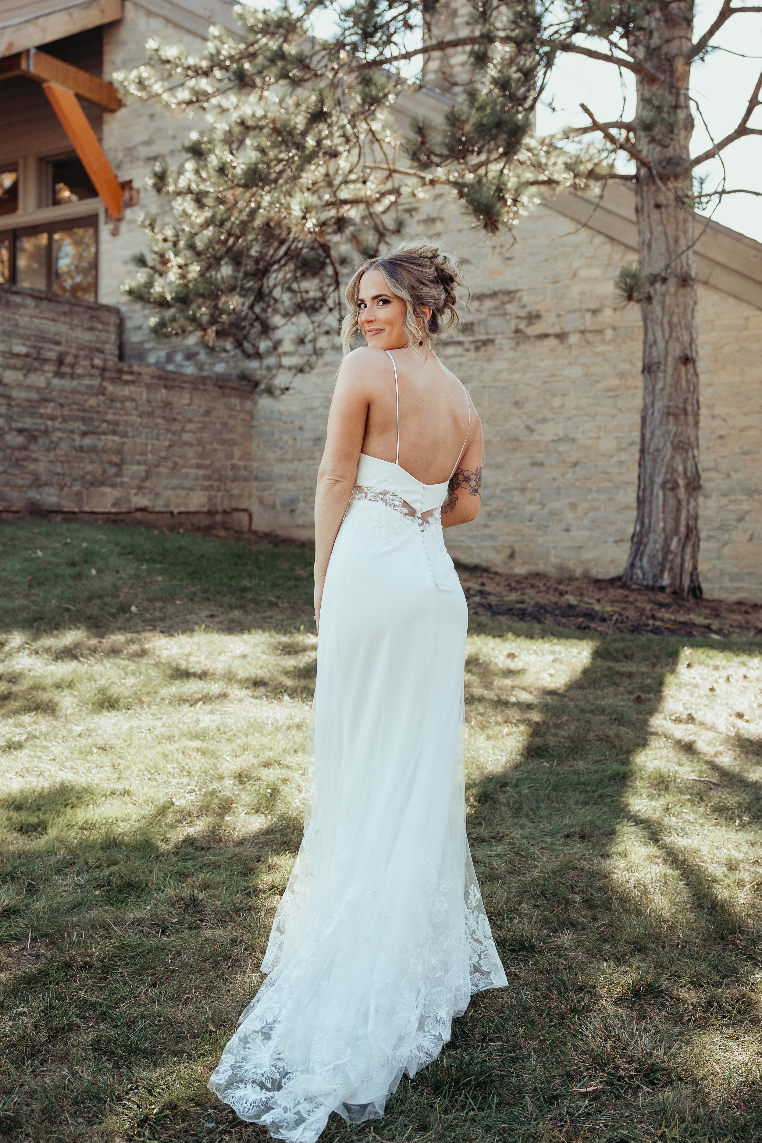 A woman in a white wedding dress standing on grass in a backyard, smiling and looking over her shoulder with trees and a stone wall in the background.