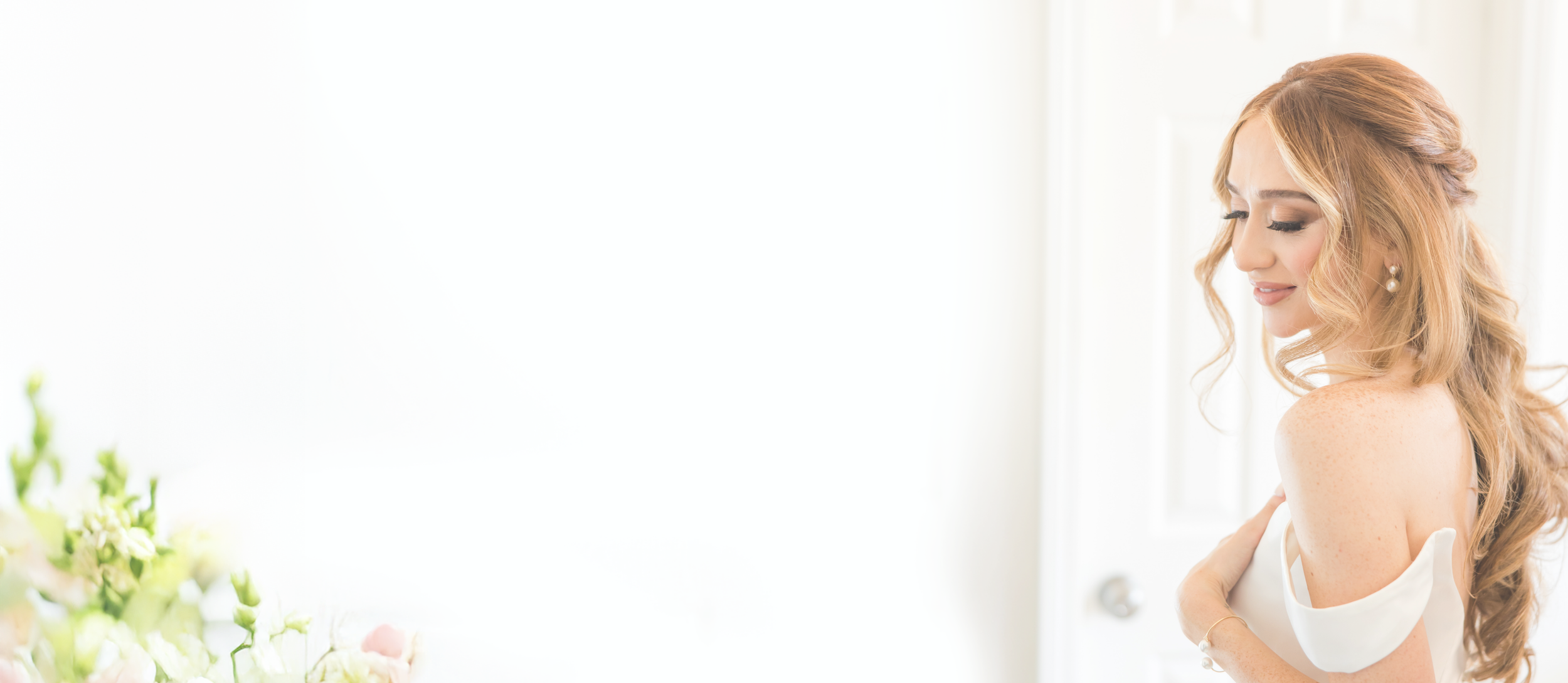 A woman with long, curly blonde hair wearing pearl earrings and a white off-the-shoulder dress, smiling softly with her eyes closed in a bright room.