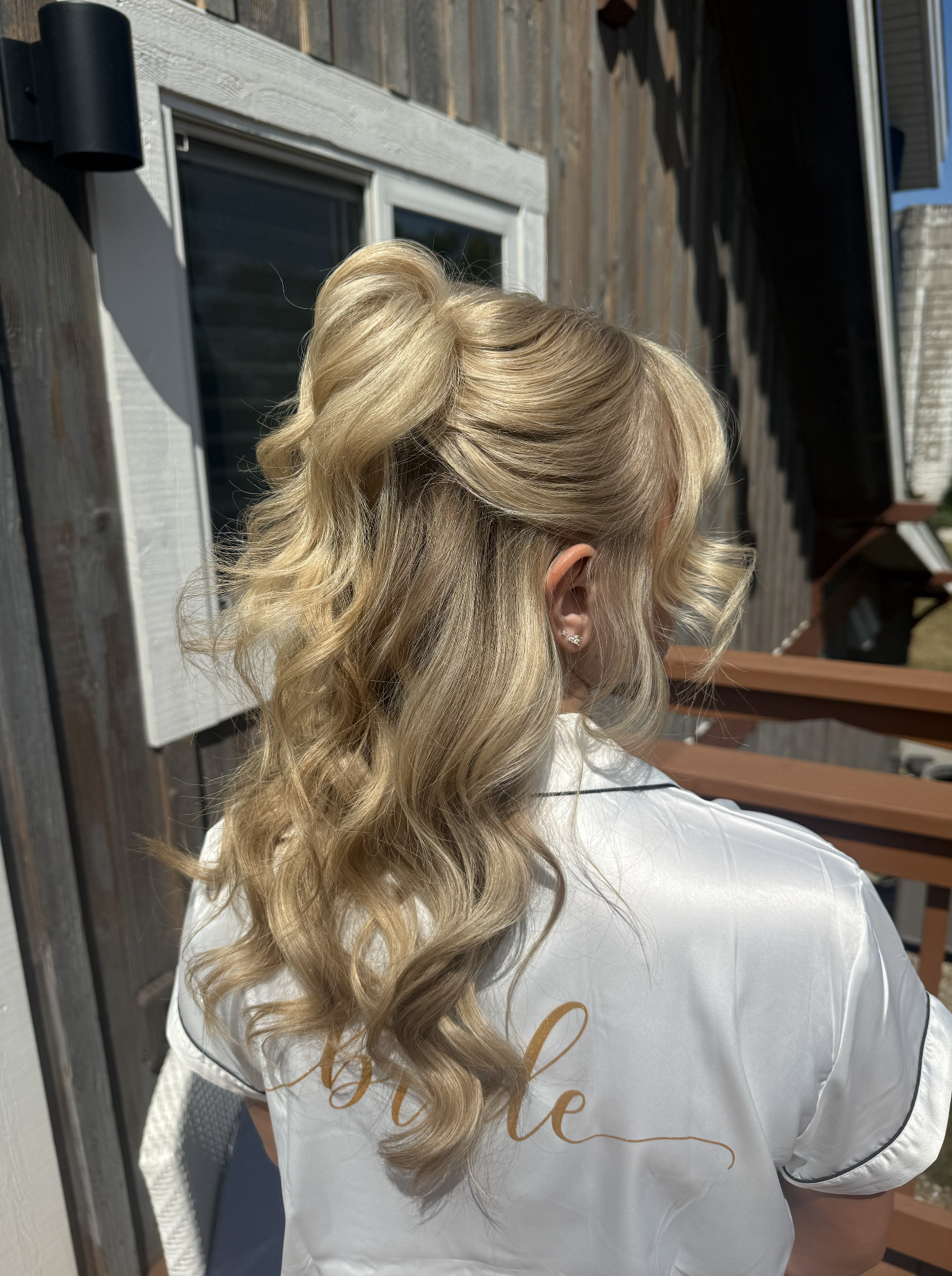 Back view of a woman with long, styled blonde hair with loose curls, standing outdoors near a wooden building, wearing a white satin robe with gold writing.