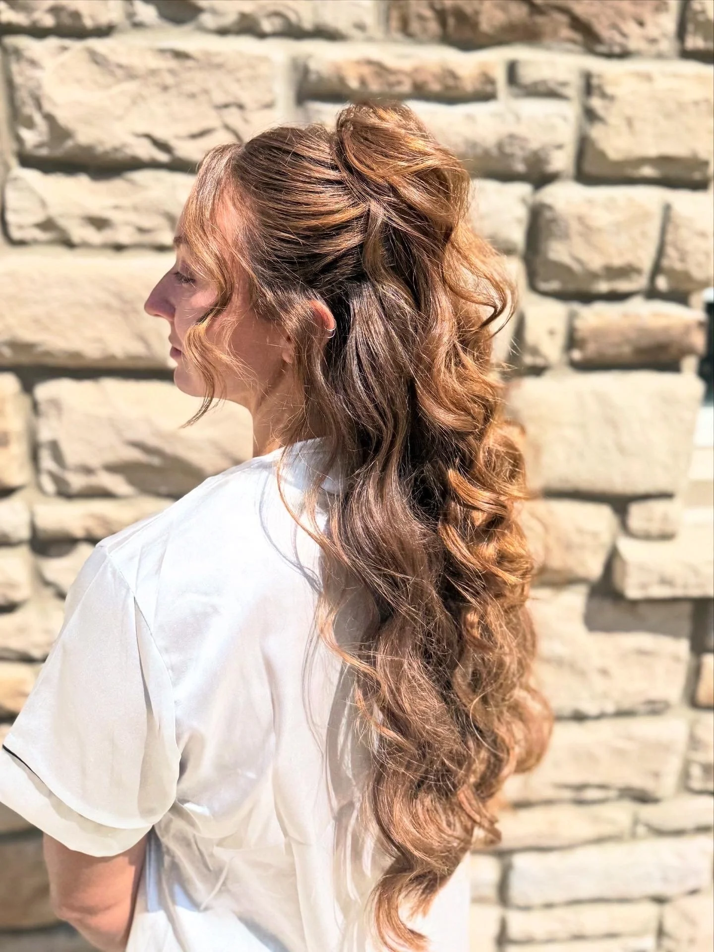 Profile of a woman with long, curly, reddish-brown hair against a stone wall, wearing a white shirt.