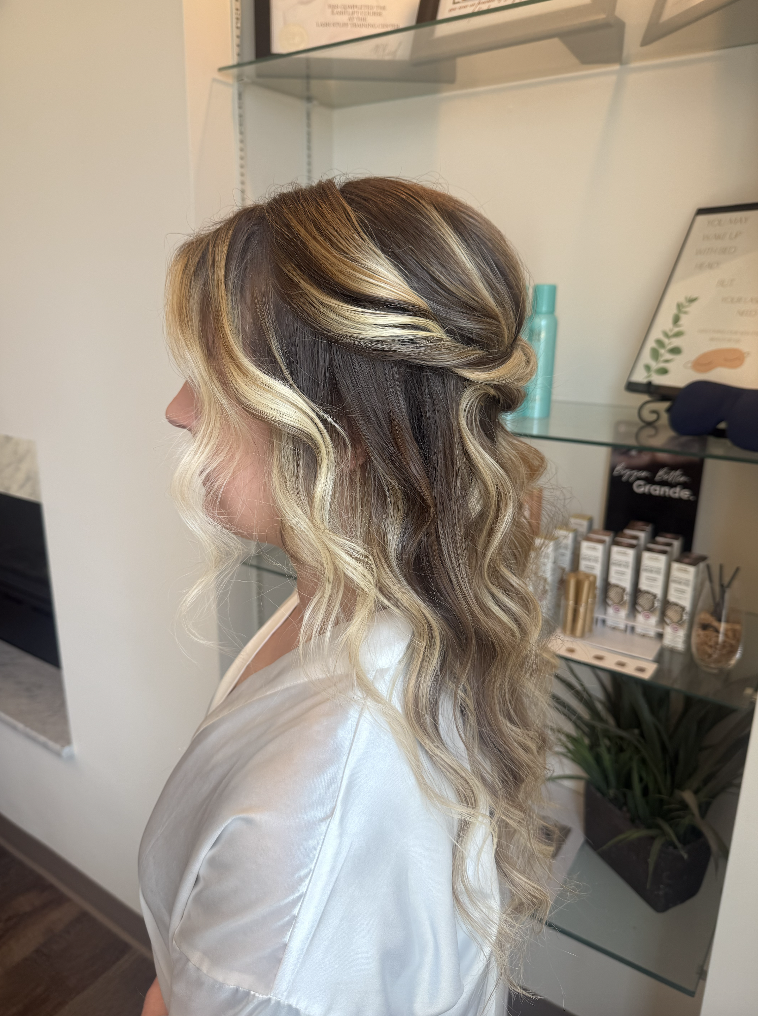 Side view of woman with long hair styled in loose curls with blonde highlights and darker roots, wearing a white silk robe in a salon.