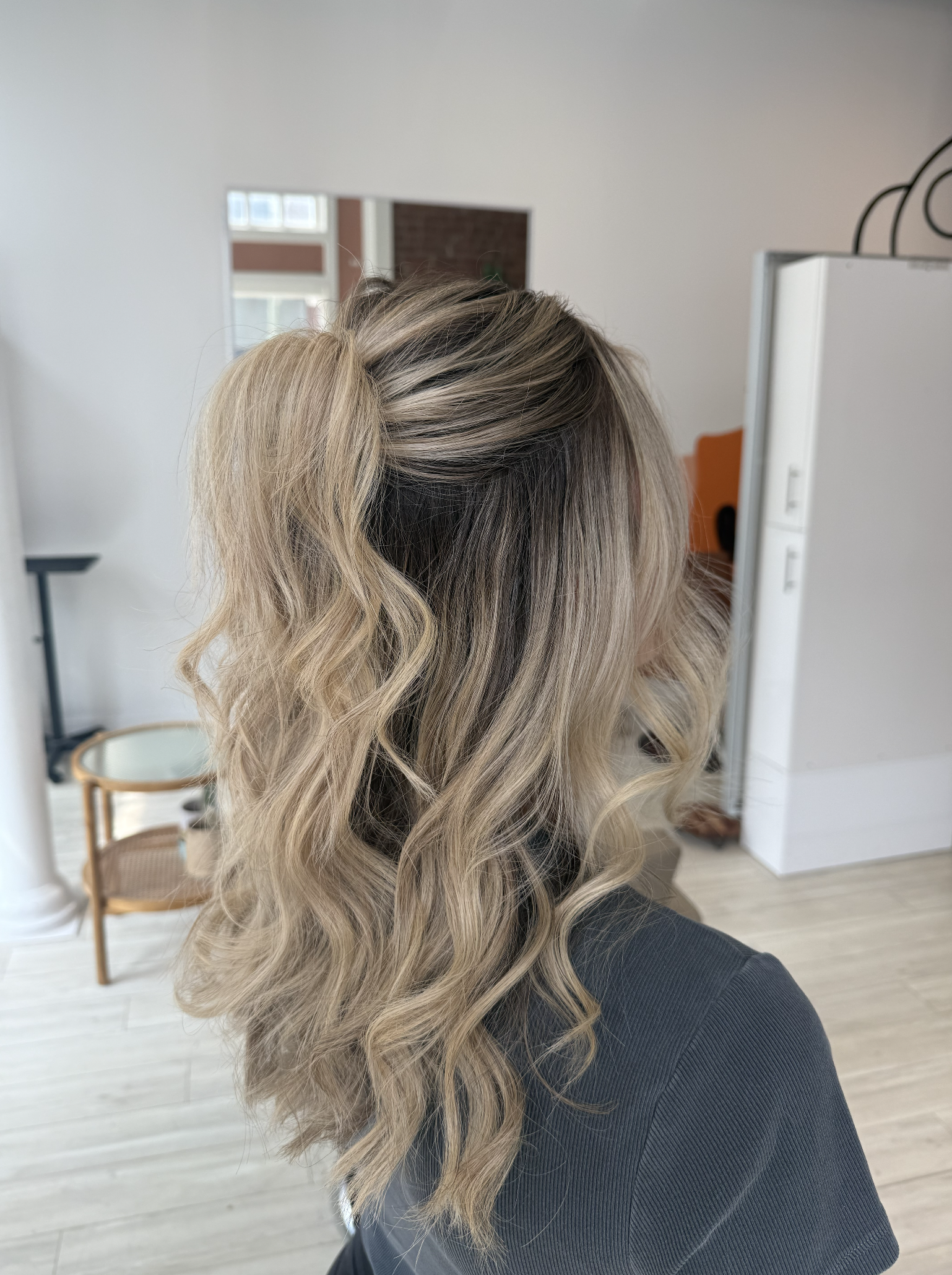 Woman with styled blonde, wavy hair in a salon or studio, seen from the back.