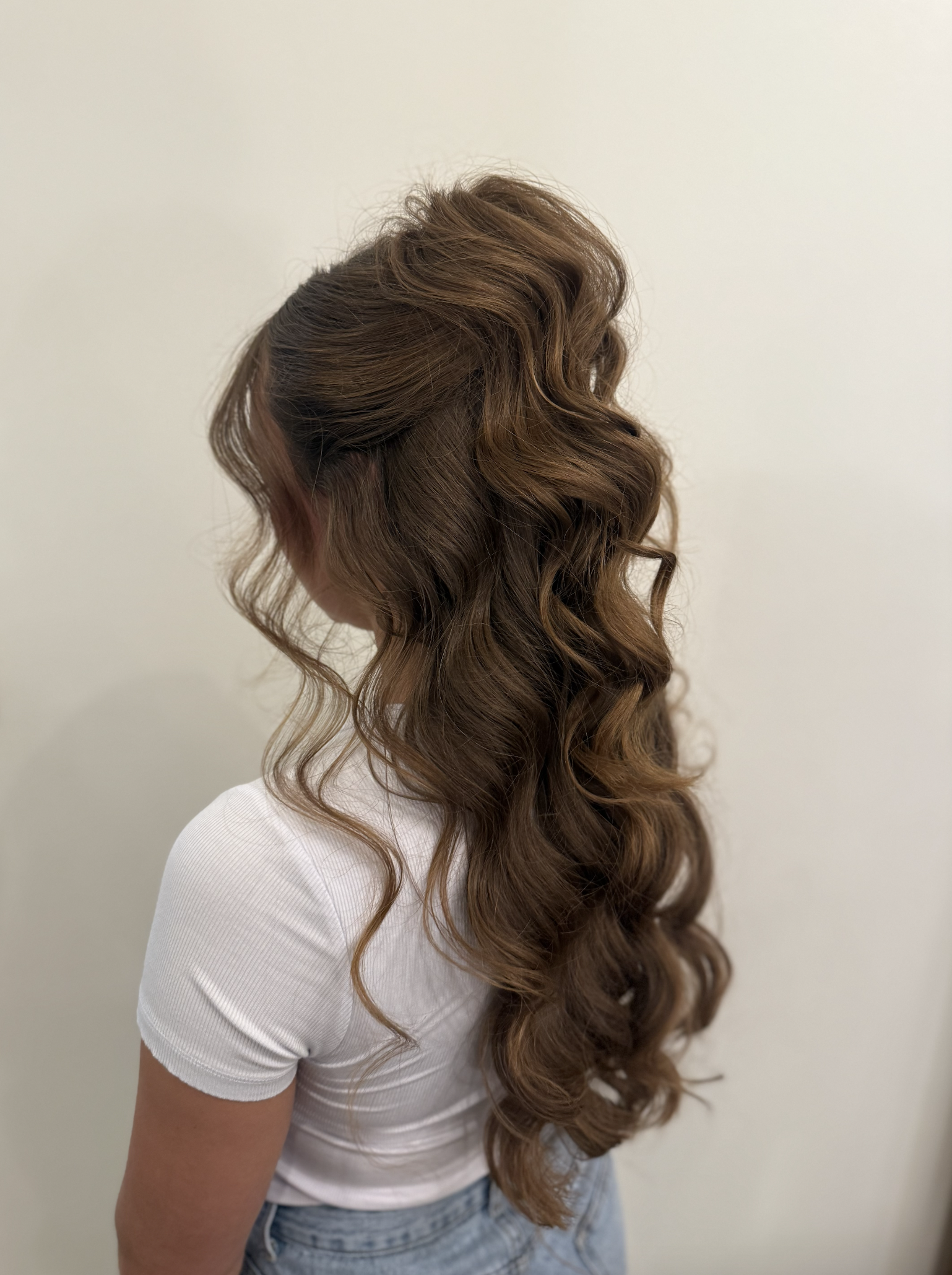 Side view of a woman with long, wavy, light brown hair, wearing a white short-sleeve shirt and blue jeans, standing against a plain light-colored background.