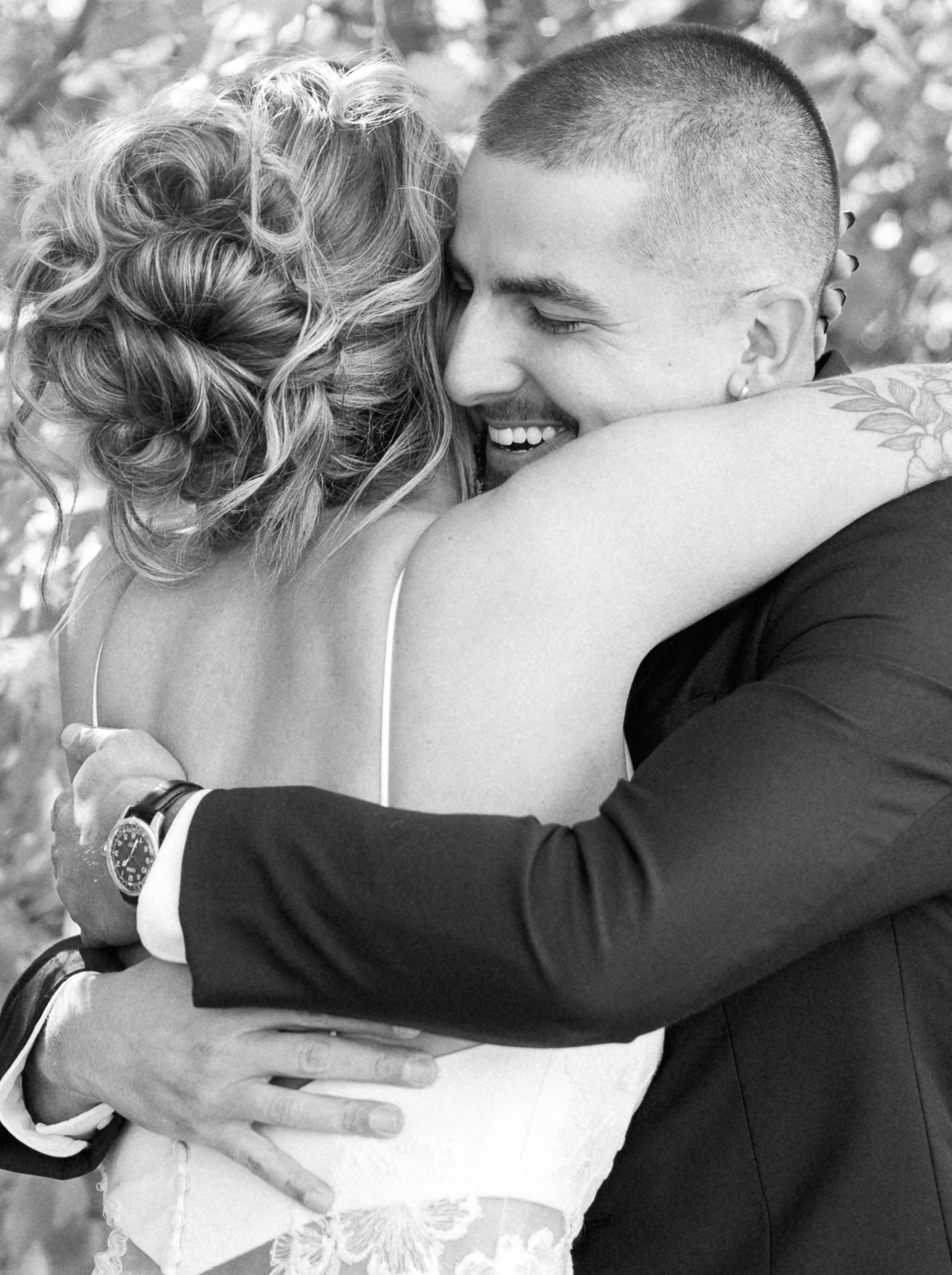A black-and-white photo of a couple hugging, smiling, with their faces close together. The woman has curly hair and is wearing a dress, while the man has a buzz cut, a tattoo on his arm, and is wearing a suit.