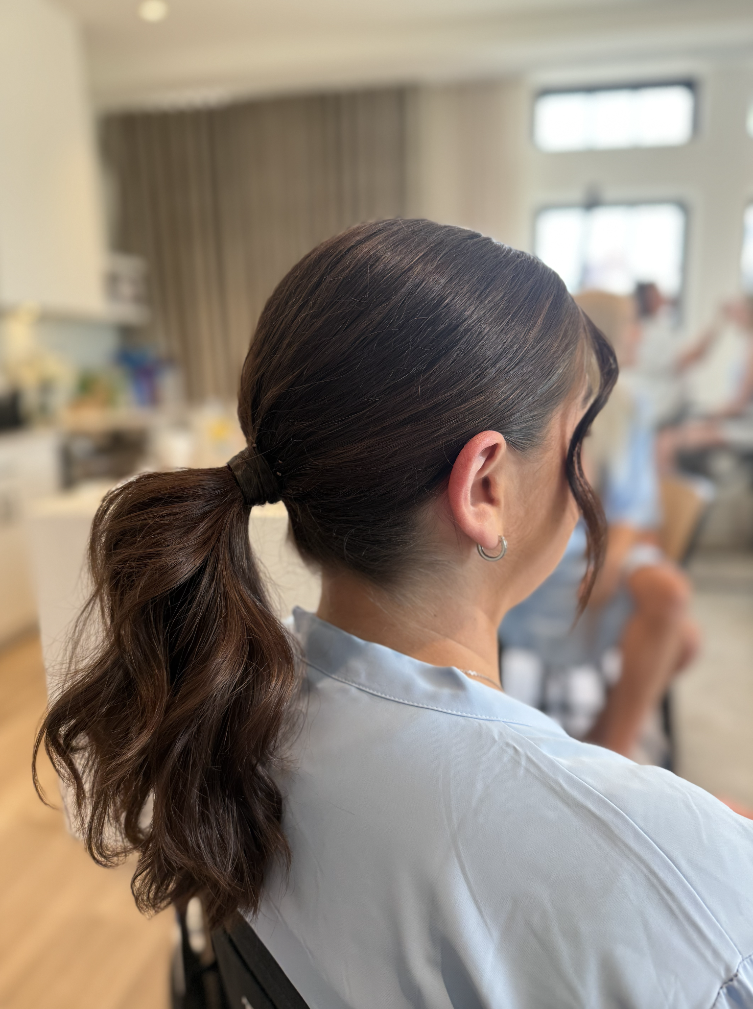 A woman with dark brown, wavy hair tied in a ponytail, wearing a silver hoop earring and a light blue top, sitting in a room with blurred background.