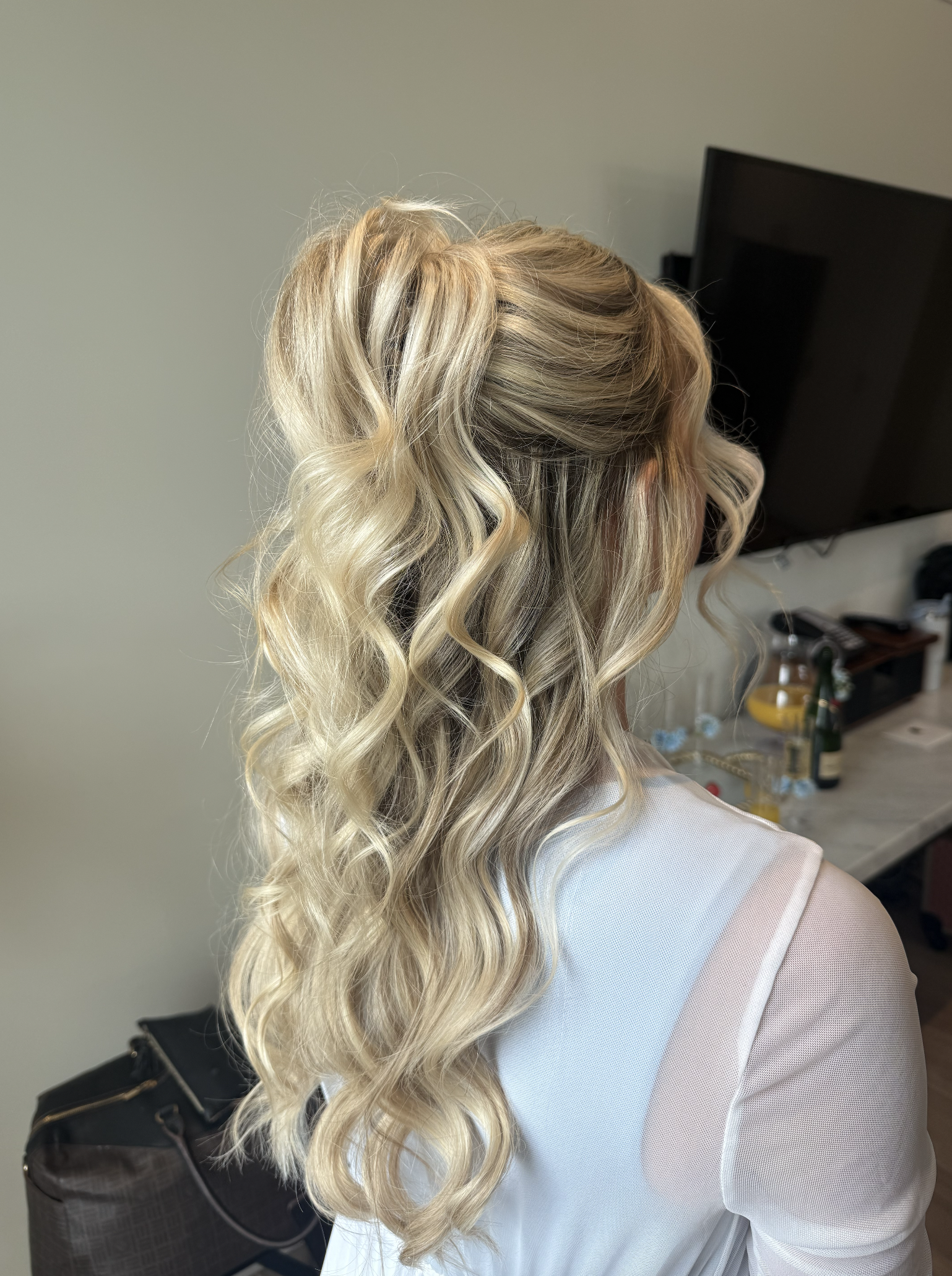 Back of a woman with long, blonde, curly hair, wearing a white sheer top, in a room with a TV and a table with drinks and decorations.
