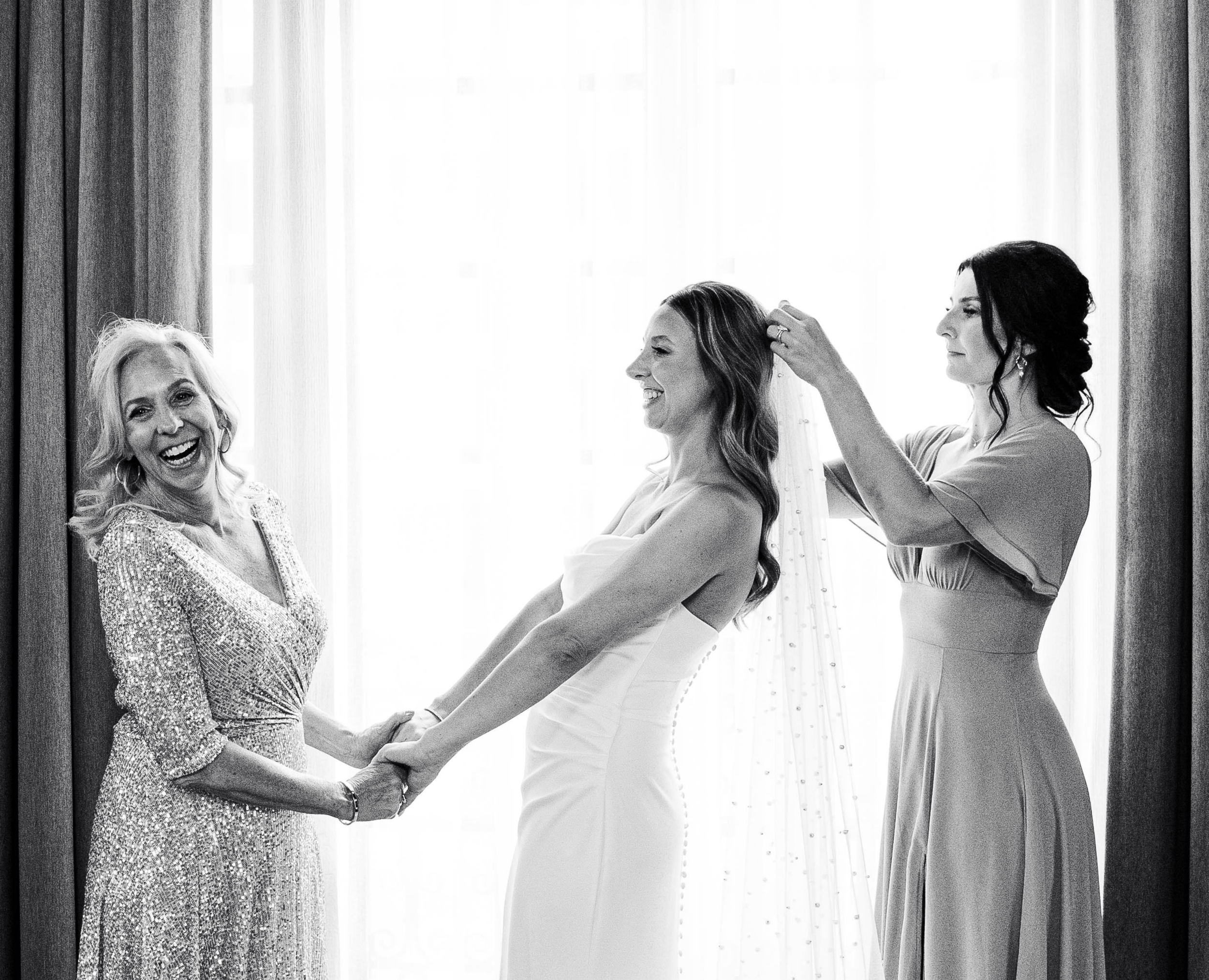 A bride holding hands with her mother and having her veil adjusted by another woman in a softly lit room.