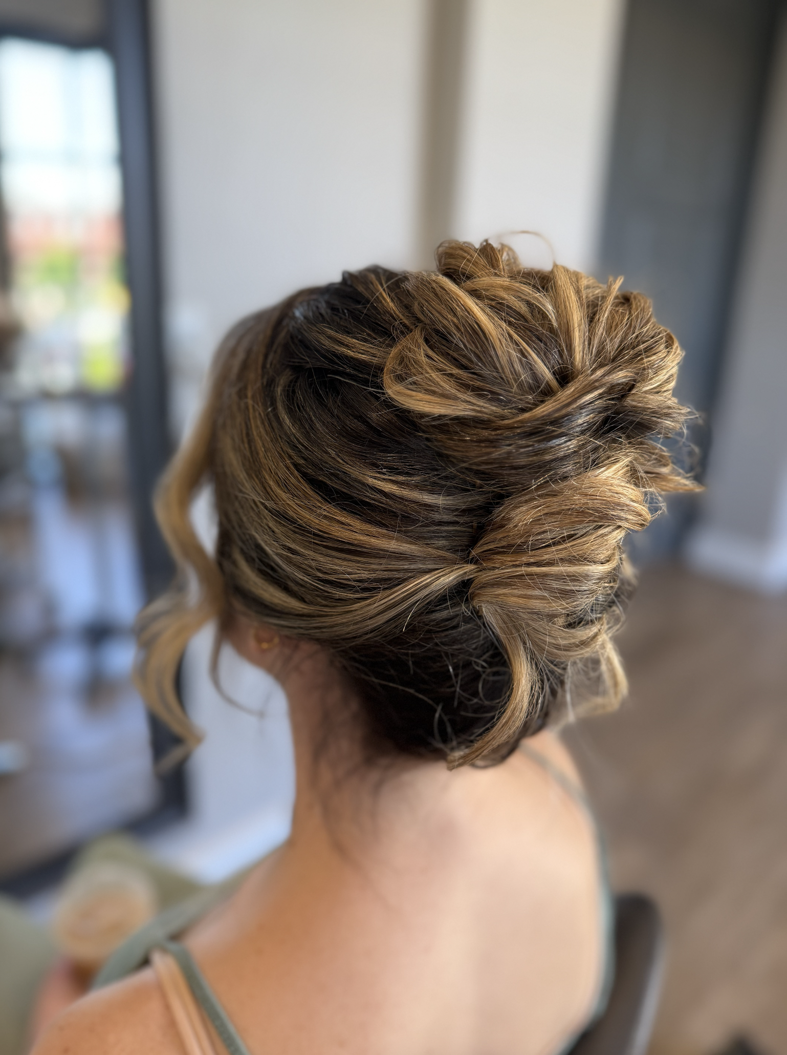 Woman with styled, layered, caramel blonde hair in an elegant updo, viewed from the back of her head.