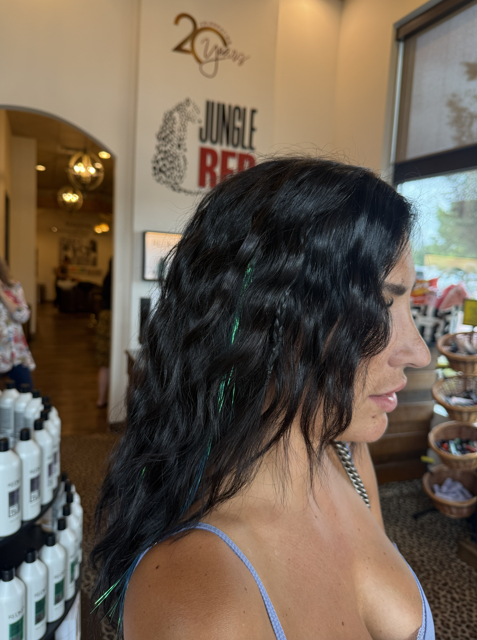 A woman with curly black hair featuring green highlights and a braid stands inside a store near a window, with shelves of beauty products nearby and a large sign on the wall that reads '20 Years, Jungle Red.'