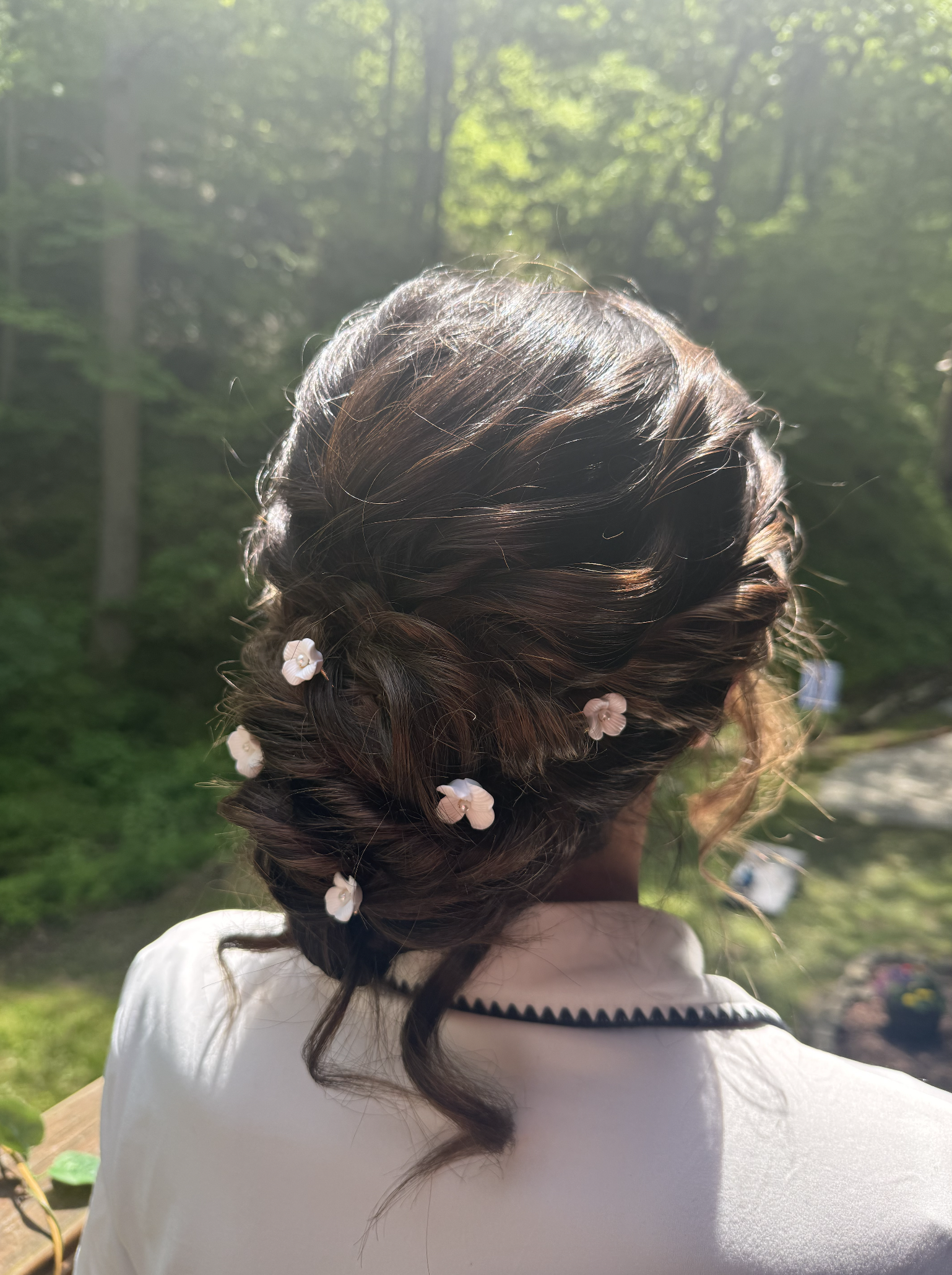 Back of a woman's head with braided hairstyle decorated with white flowers, outdoors with trees and sunlight in the background.