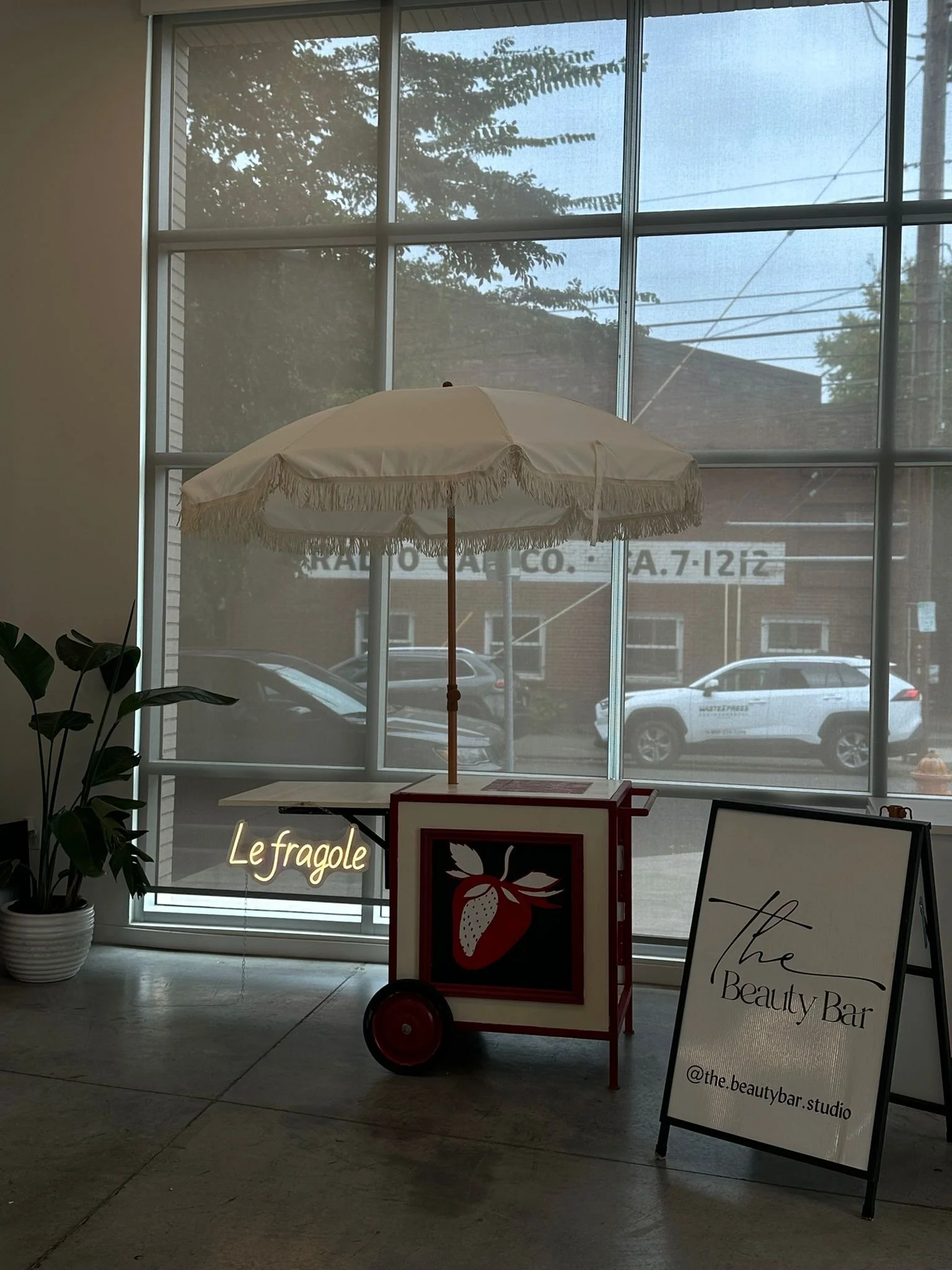 A mobile strawberry smoothie cart with a white umbrella inside a store near a large window, with a potted plant on the left and a sign reading 'The Beauty Bar' on the right.