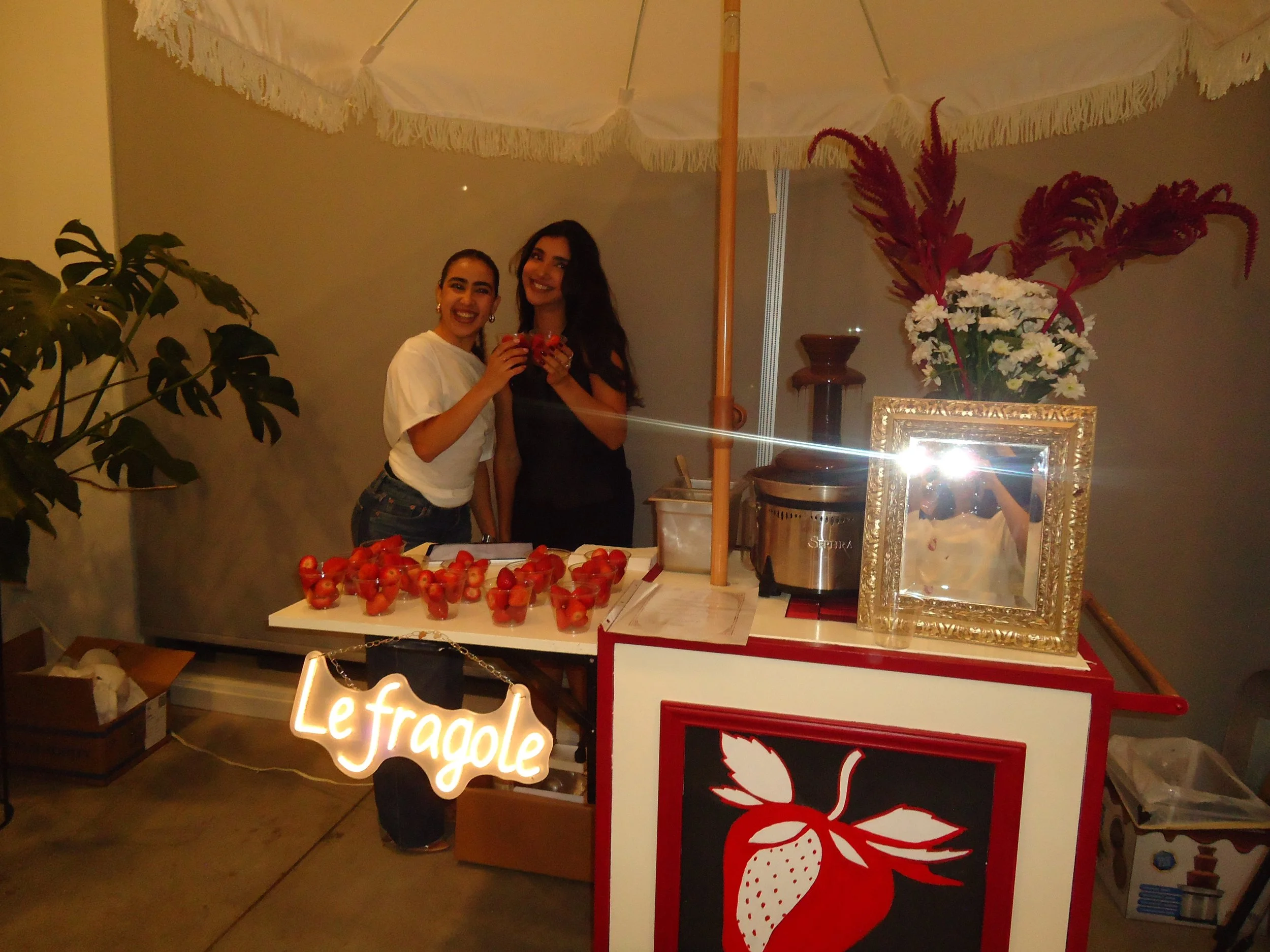 Two women stand behind a strawberry stand, smiling and holding strawberries. The stand is decorated with a sign that says 'Le Fragole' and a large strawberry graphic. There are fresh strawberries on the table, a flower arrangement behind them, and a