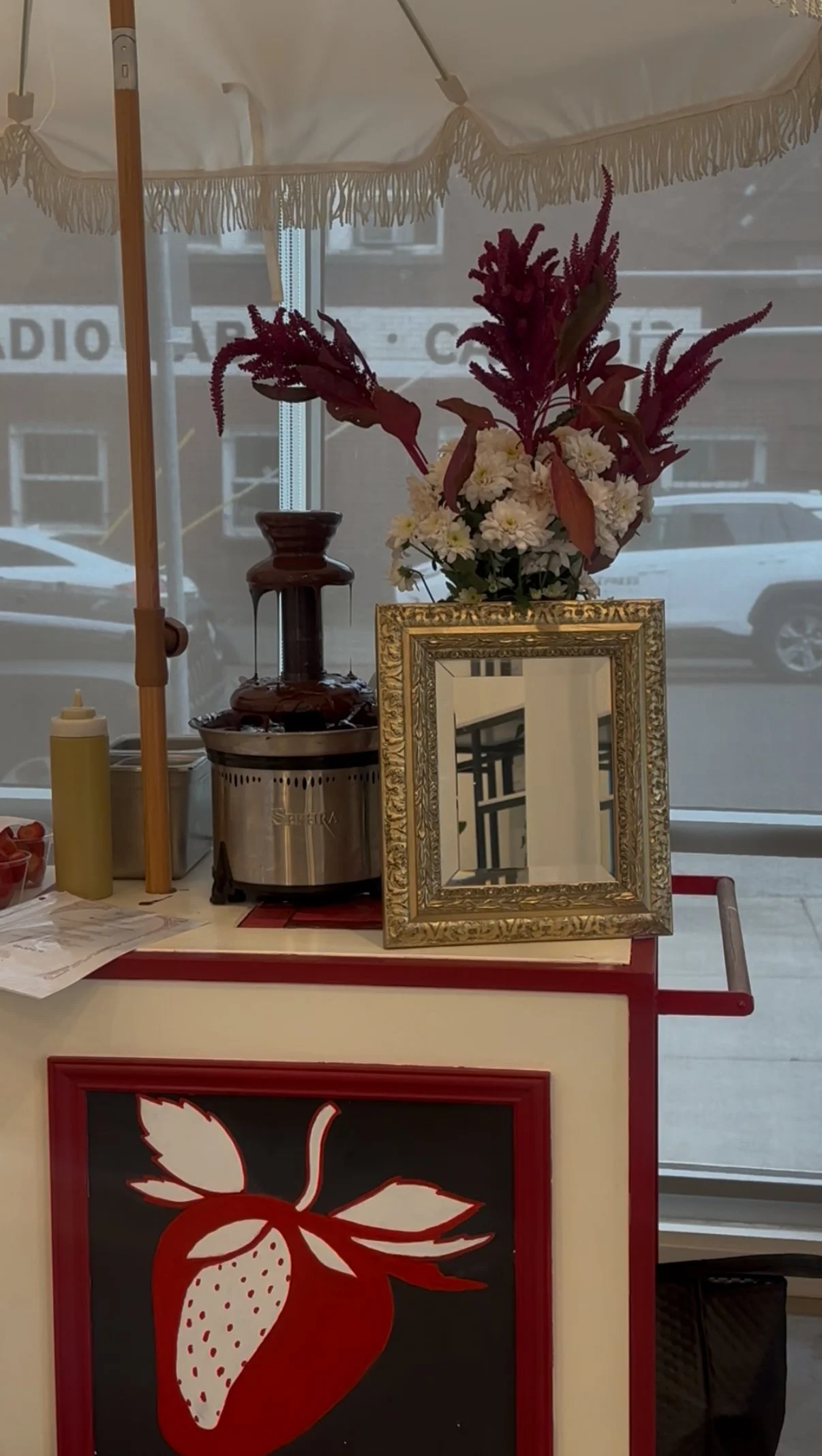 A flower arrangement with white flowers and purple leaves in a gold ornate frame, a chocolate fountain, and a yellow squeeze bottle on a table with an illustration of a strawberry on the front. The background shows a window with a parked car outside.