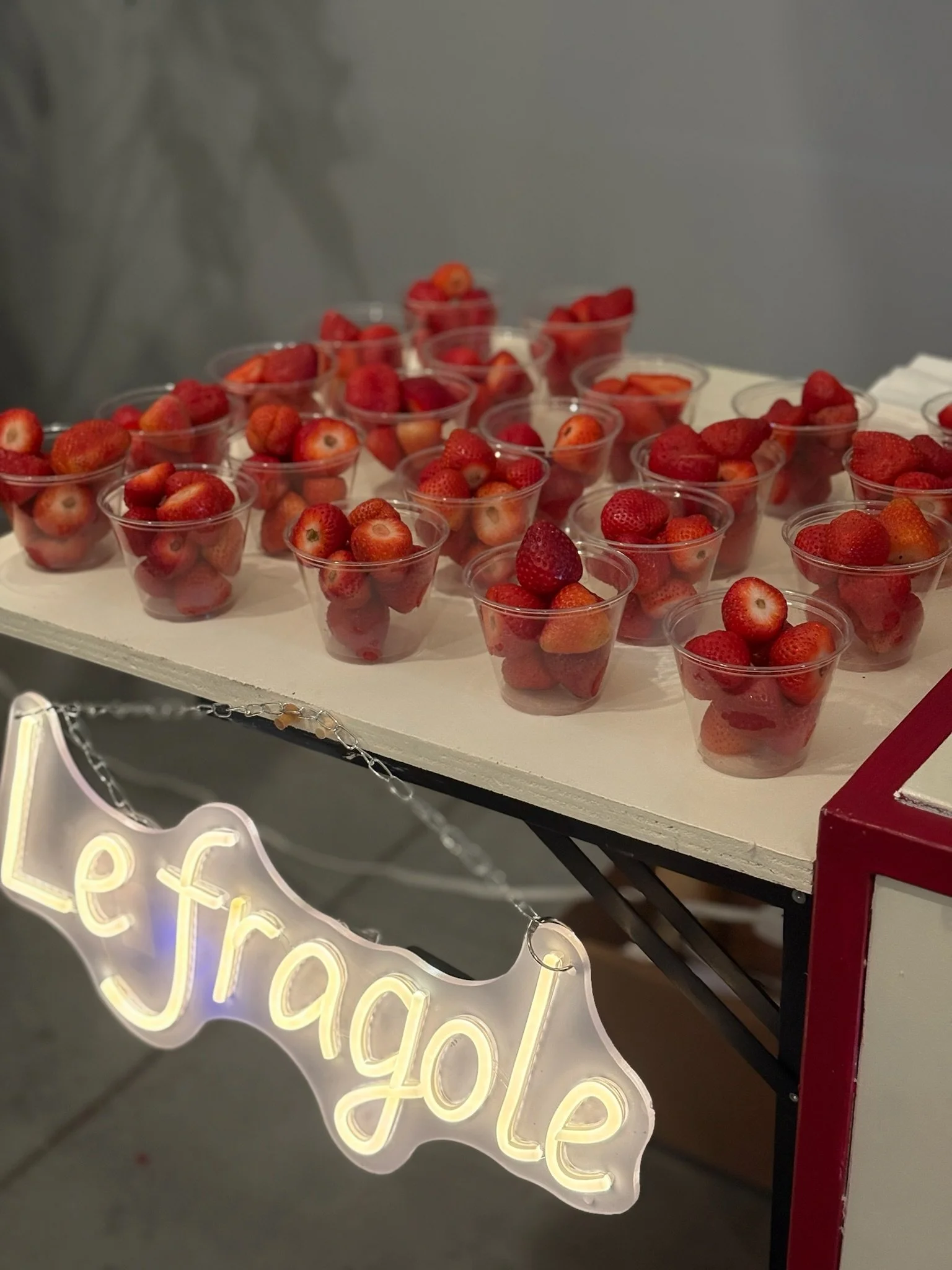 Small cups filled with strawberries labeled as a strawberry sale stand with a neon sign that reads "Le fraise".