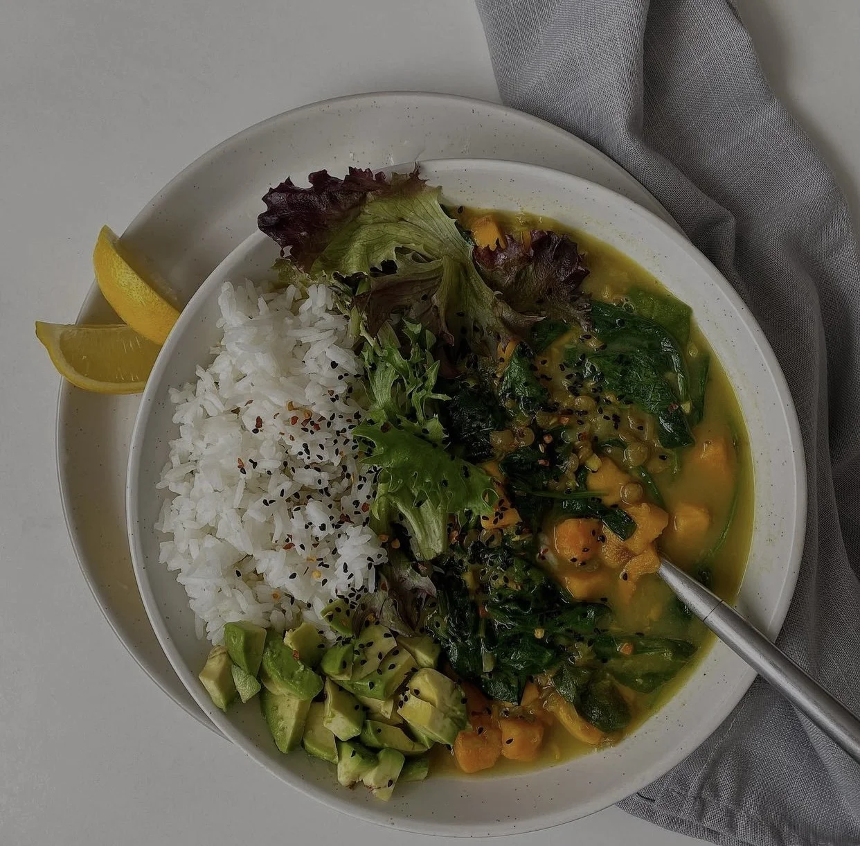 A bowl of rice with sesame seeds, mixed greens, and a vegetable curry, served with lemon wedges on the side.