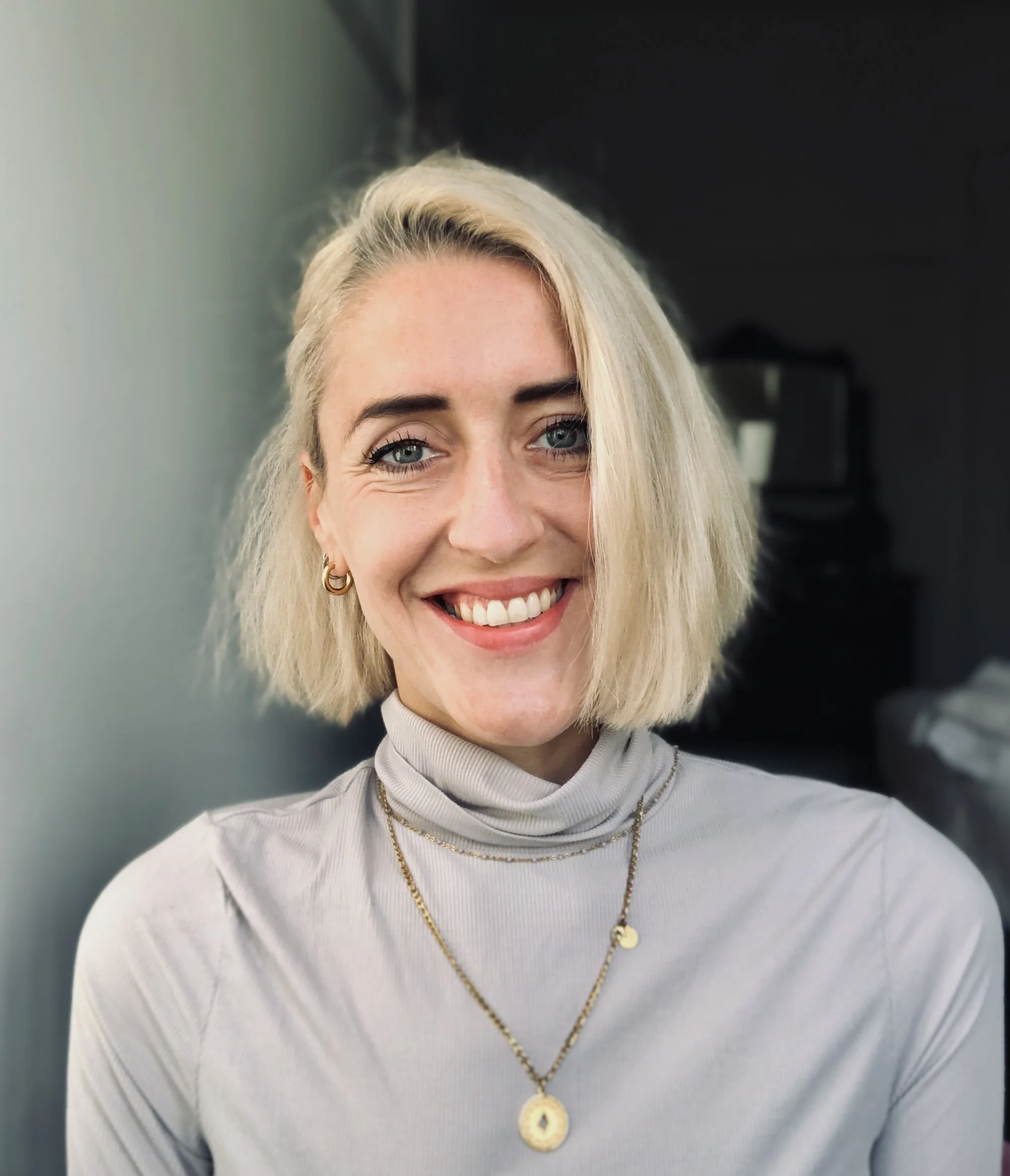 Smiling blonde woman with turquoise eyes, wearing gold earrings and necklaces, in a gray turtleneck shirt, a dark room in the background.